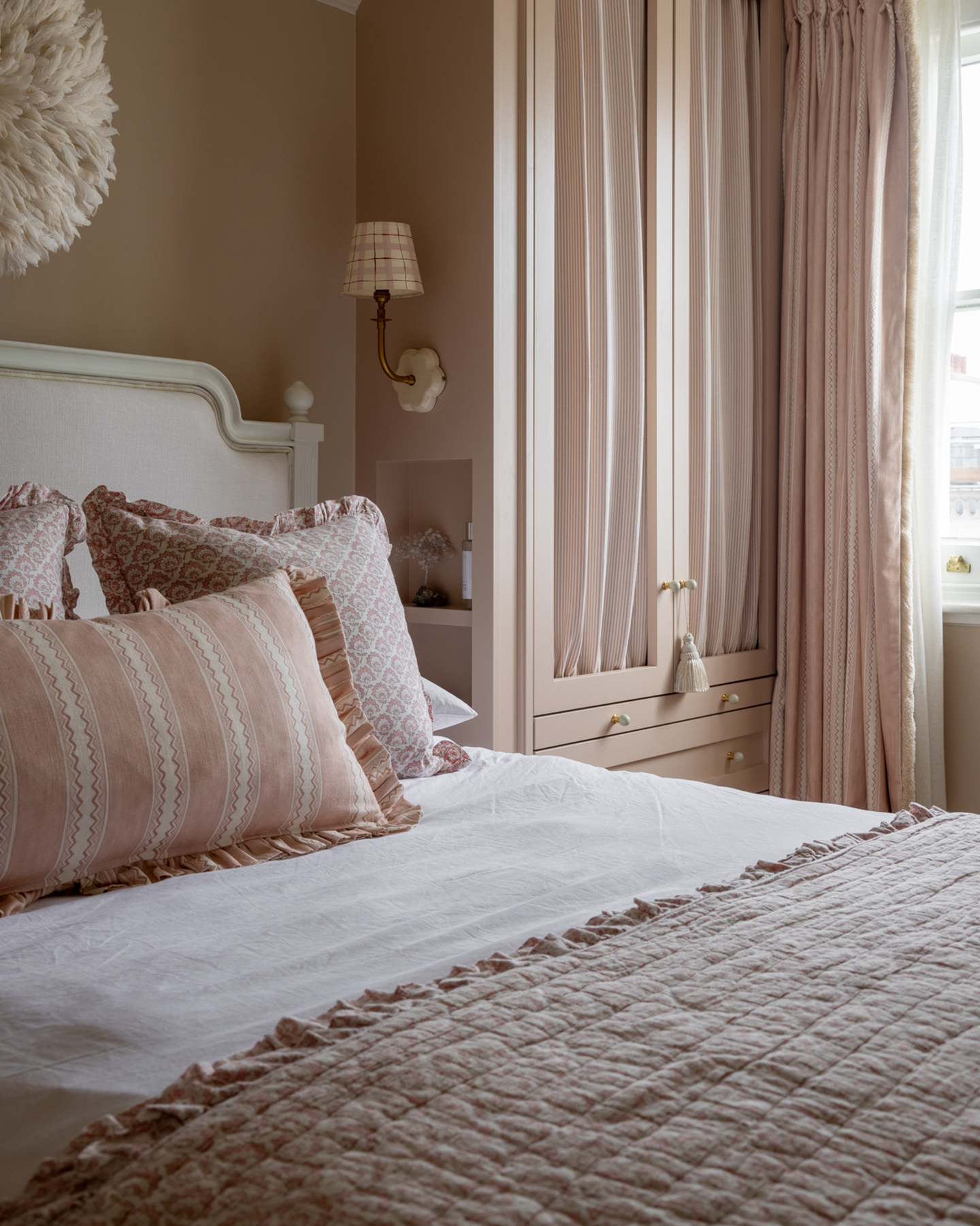 a small bedroom with pale pink walls and a built in wardrobe, a bed with white bedding and pale pink and white floral cushions and bedspread