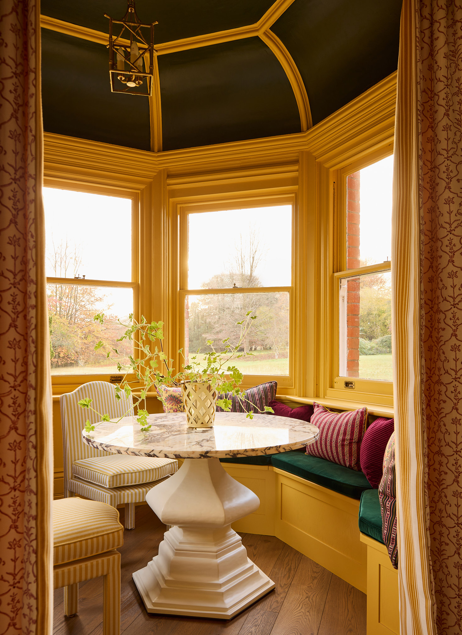 Sunny bay window breakfast nook with warm yellow millwork, a green velvet banquette, round marble pedestal table, and striped chairs