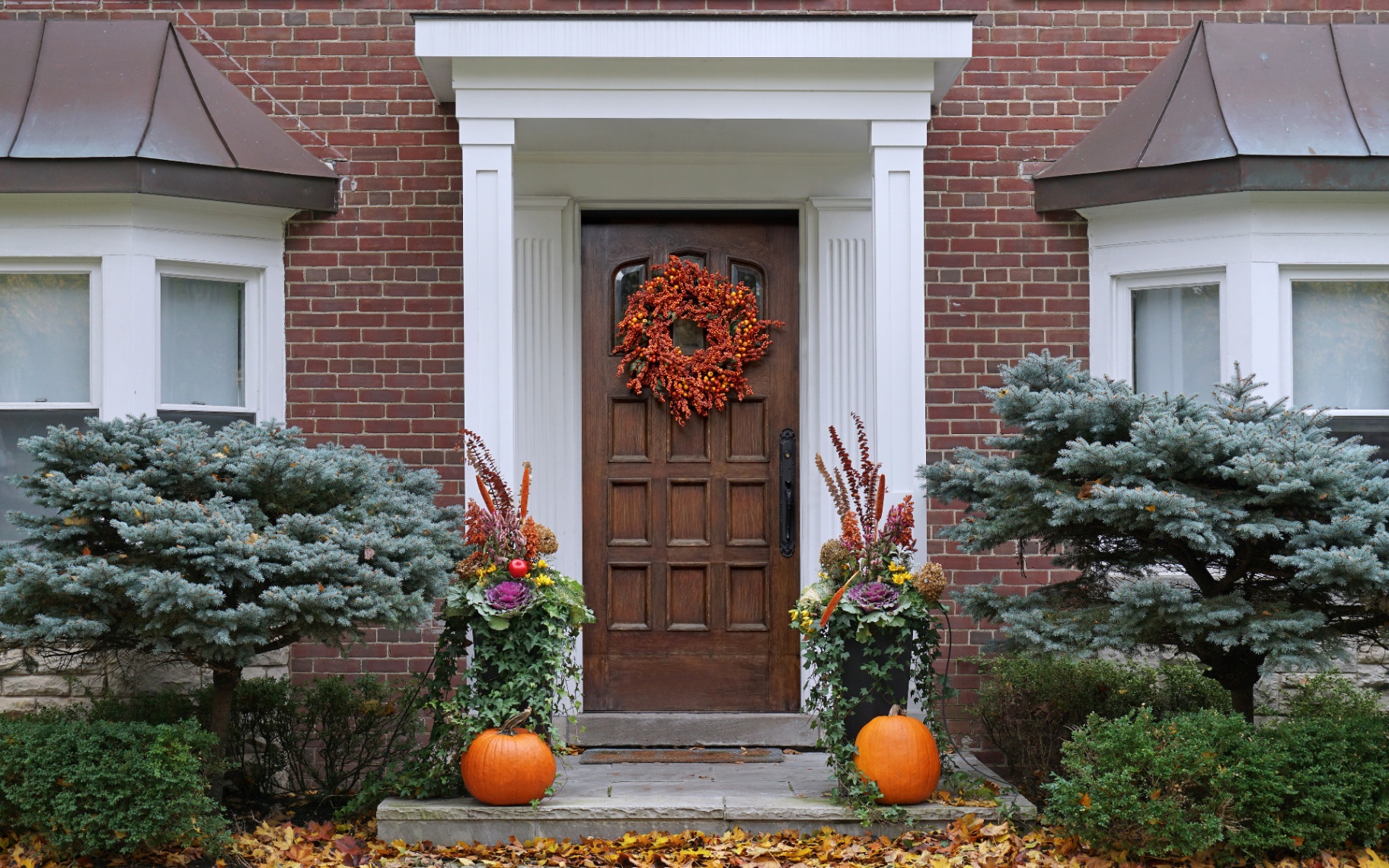 the outside of a red brick house with a dark brown front door and an autumn wreath and pumpkins on steps