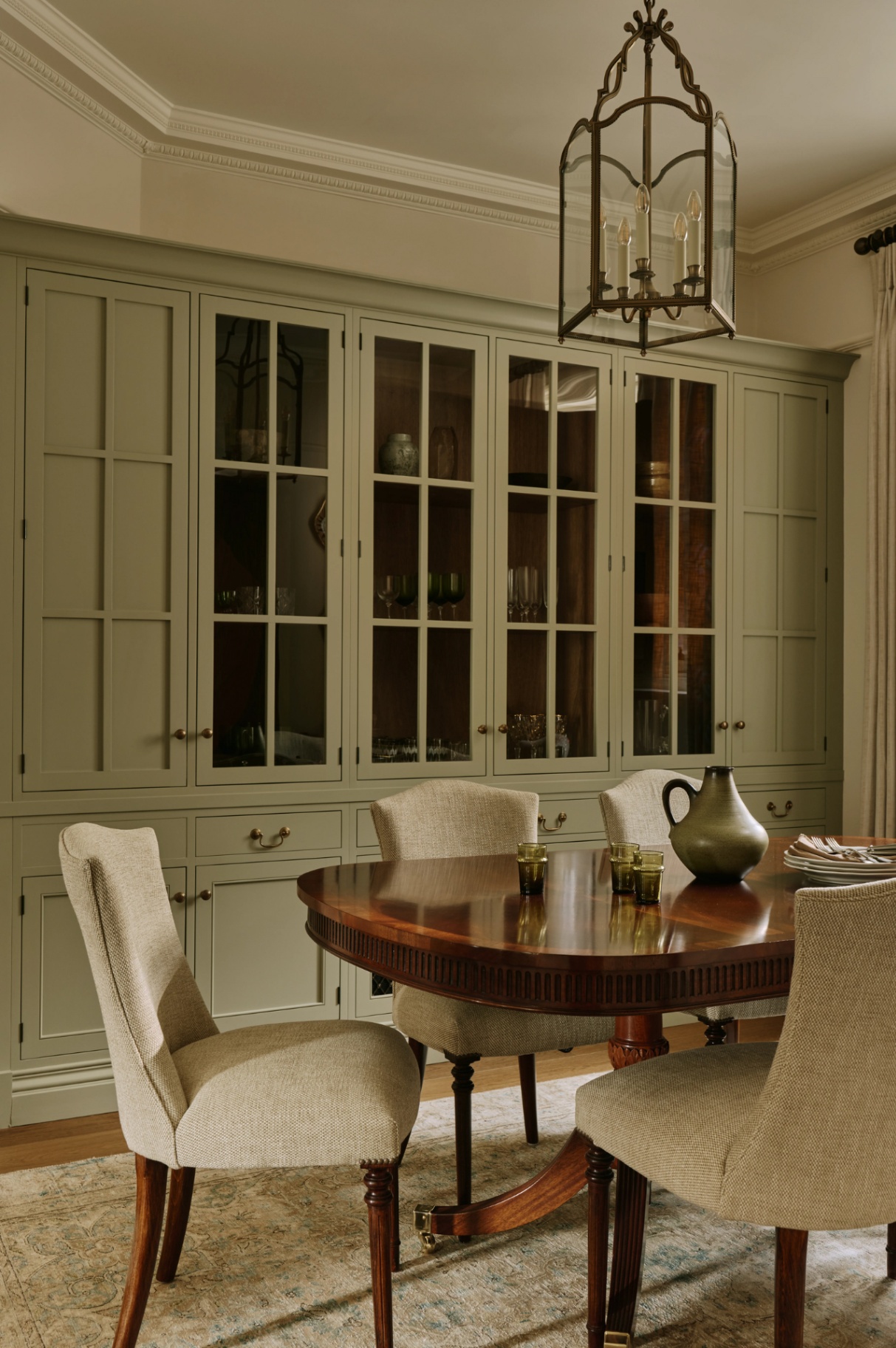 A dining room with half a large wooden table visible, surrounded by upholstered dining chairs and with a pendant lantern hanging above. On the table is a jug, glasses and plates with napkins and cutlery. A built-in cupboard runs along the back wall