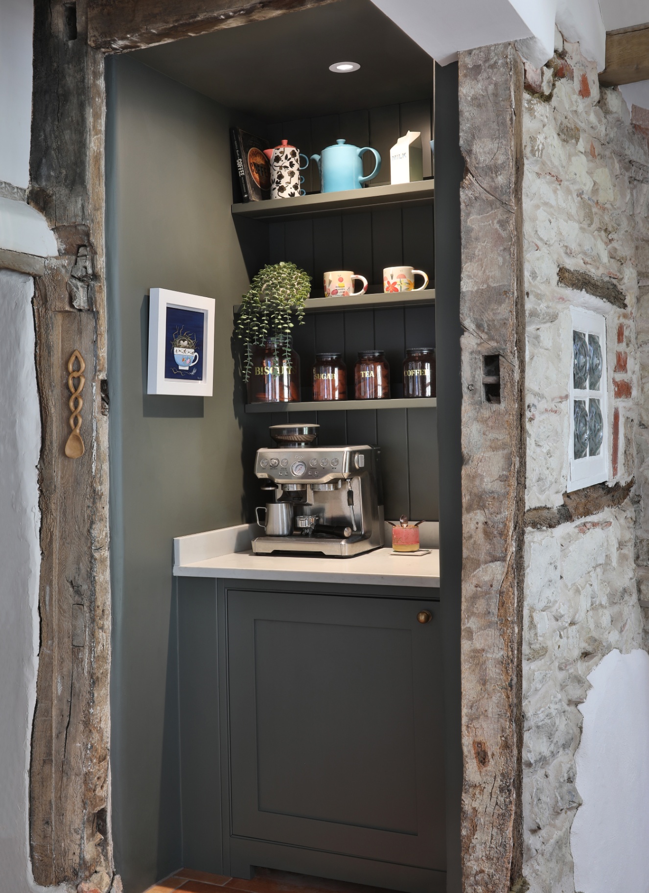 Cozy architectural alcove coffee nook framed by rustic stone and exposed timber beams, featuring deep grey painted cabinetry, open shelving for canisters and artwork, and an espresso machine