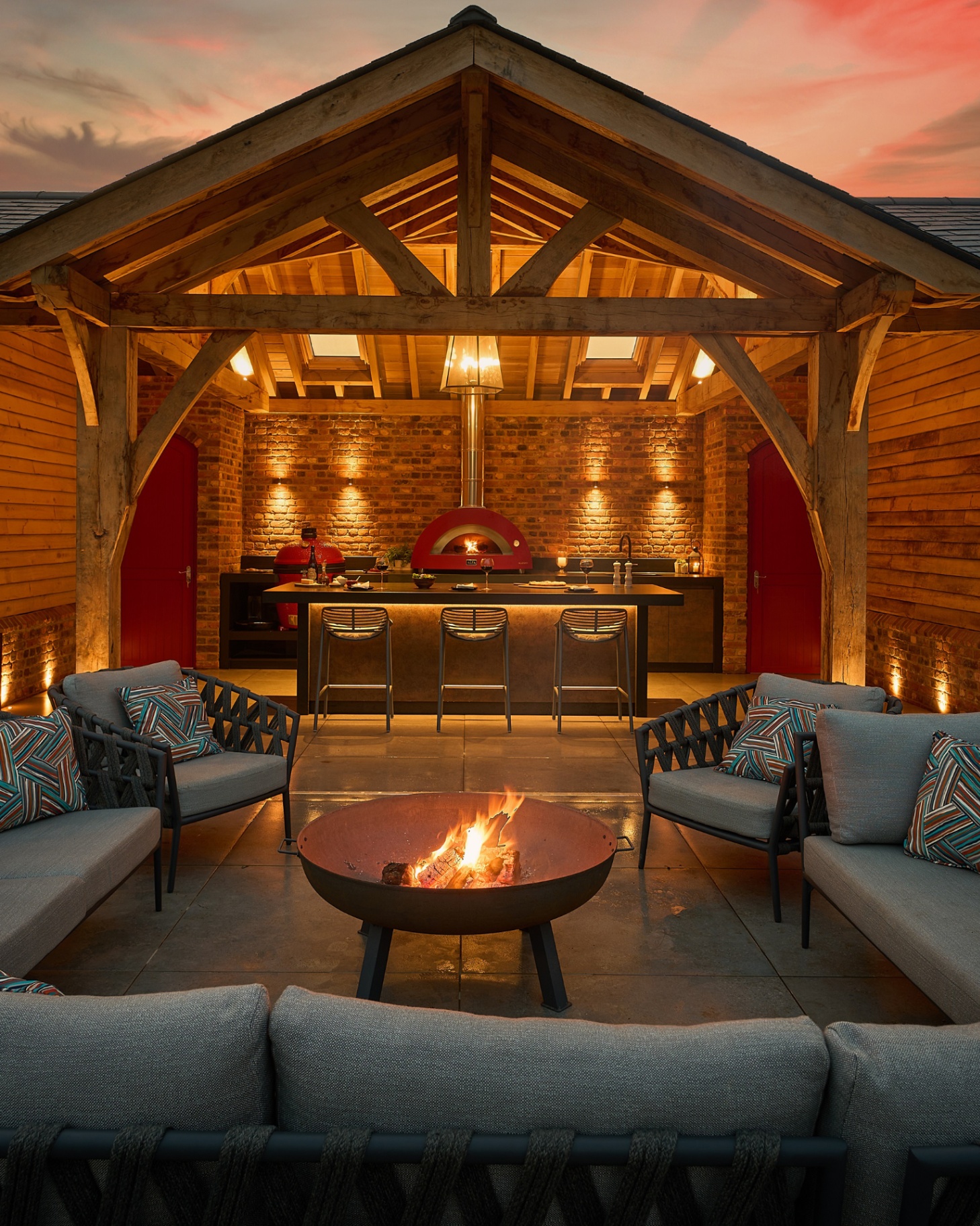 an outdoor space with an l shaped garden sofa surrounding a black bowl firepit, with an outdoor kitchen and bar area under a large wooden covering
