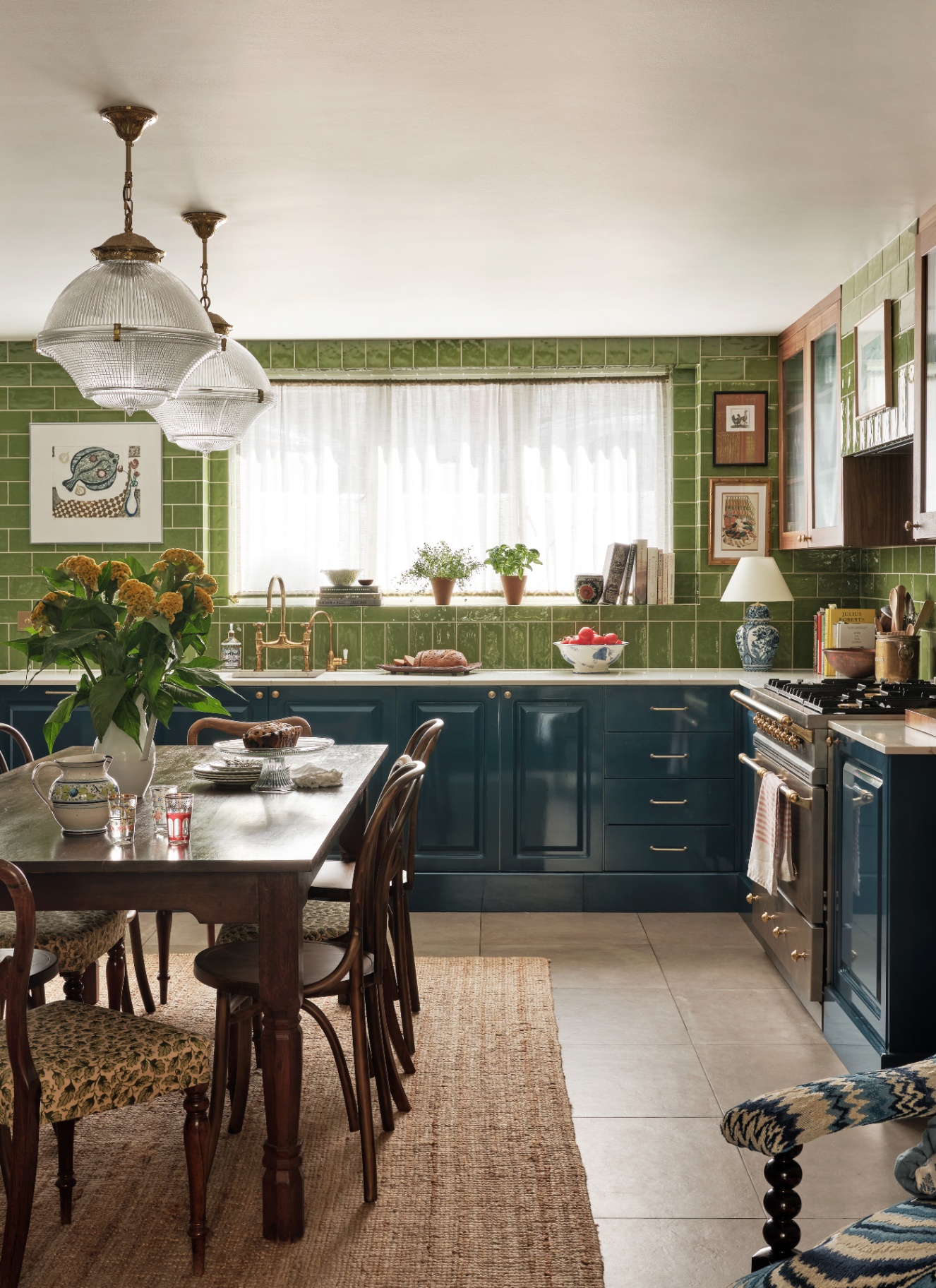 a kitchen with all over green tiles on the walls, dark blue cabinetry, two large glass pendant above a dining table and stone flooring