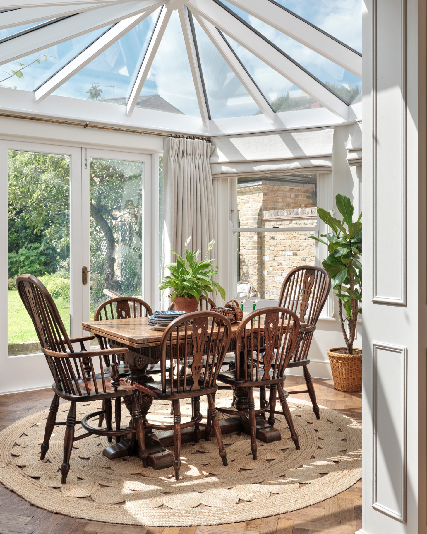 a conservatory  with grey curtains, a large mahogony dining table with spindle dining chairs and a round jute rug underneath