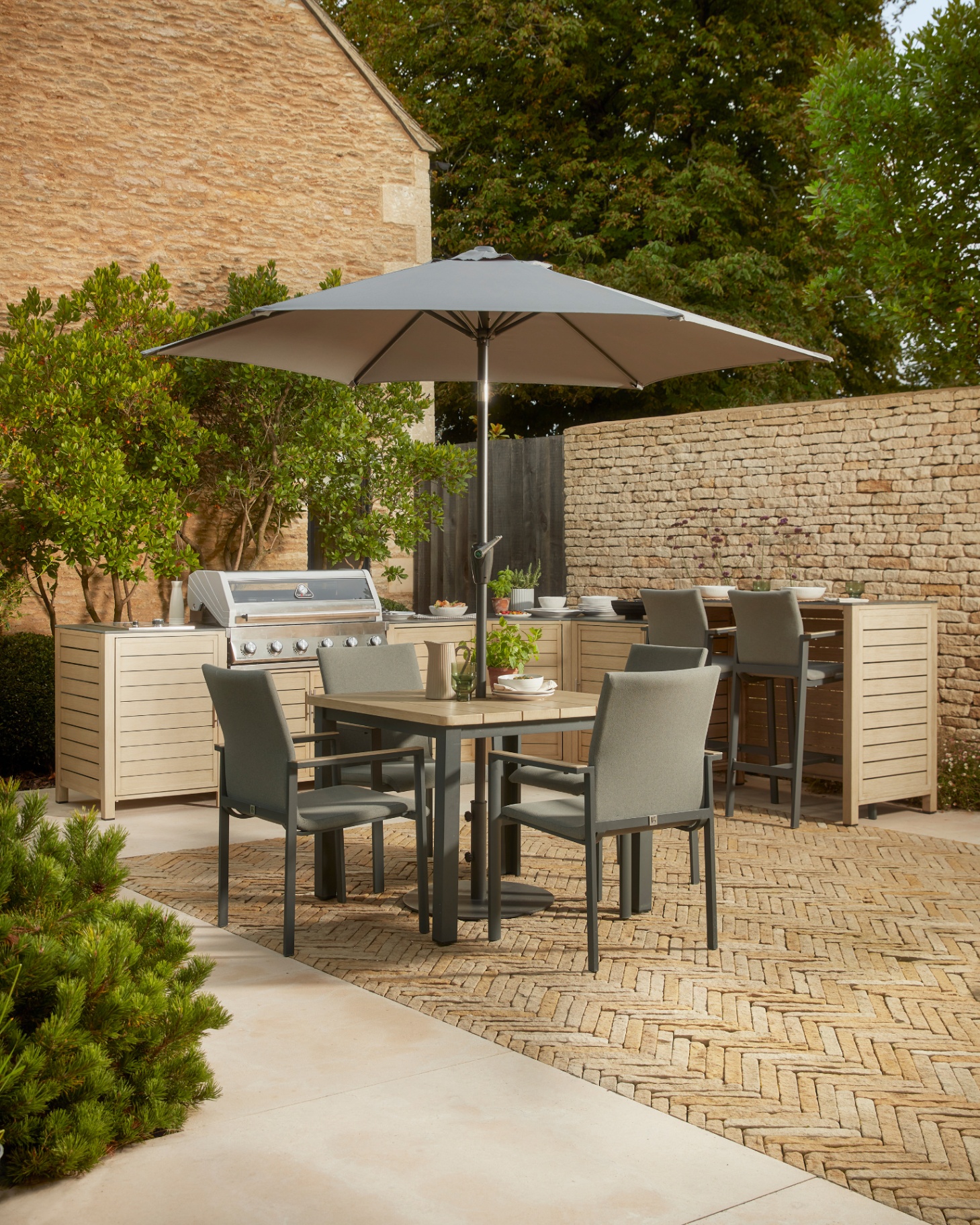 a garden with an outdoor kitchen area with a cotswold stone brick wall and trees behind it and a square outdoor dining table and chairs in front of it with a parasol