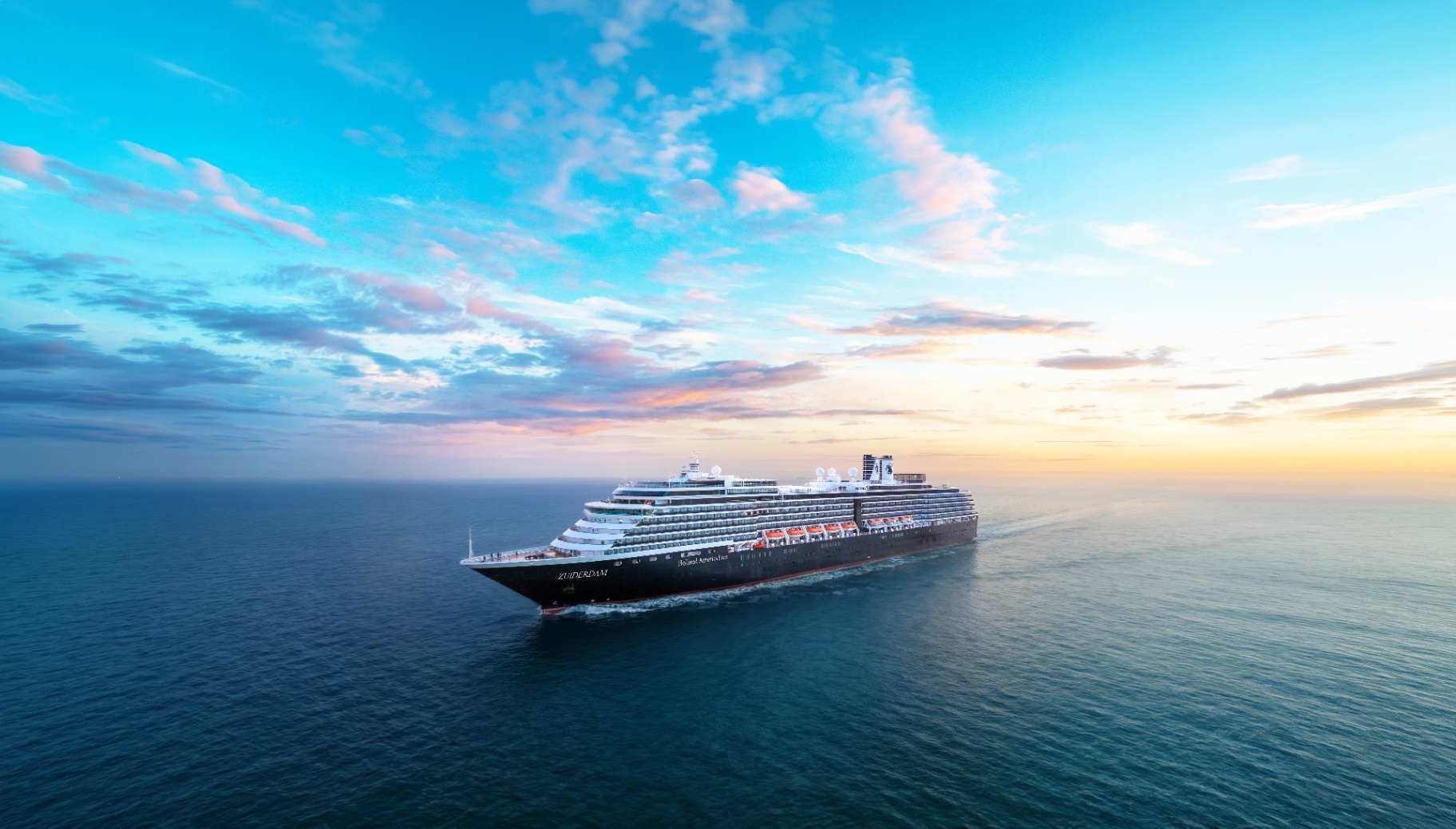 Aerial view of the cruise ship Zuiderdam sailing through open blue ocean waters under a vast sky with light, colorful clouds at sunset