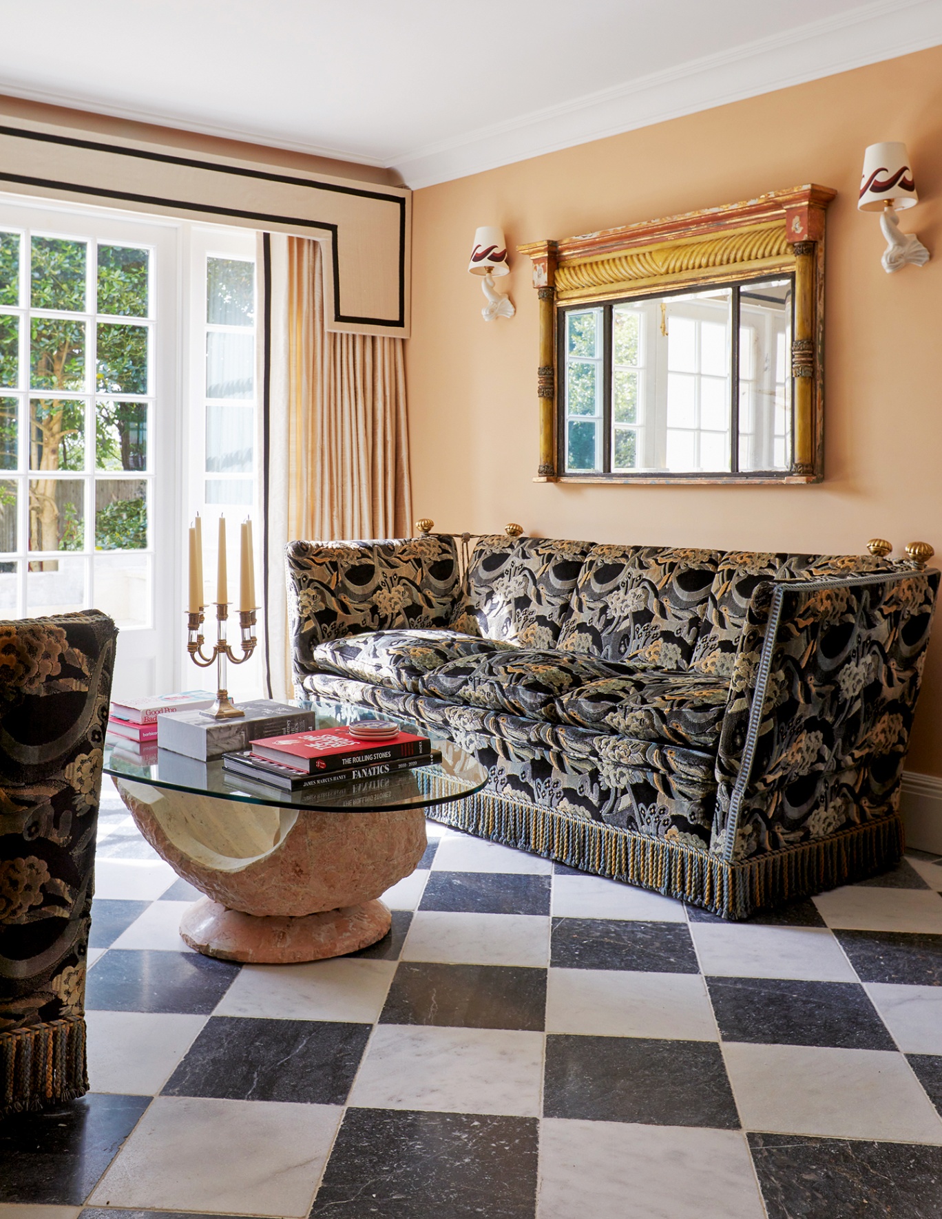 A pair of statement Knole sofas flank a sculptural stone-and-glass coffee table, atop a black and white checkerboard floor. French doors are dressed with neutral curtains and a graphic pelmet with a black line border
