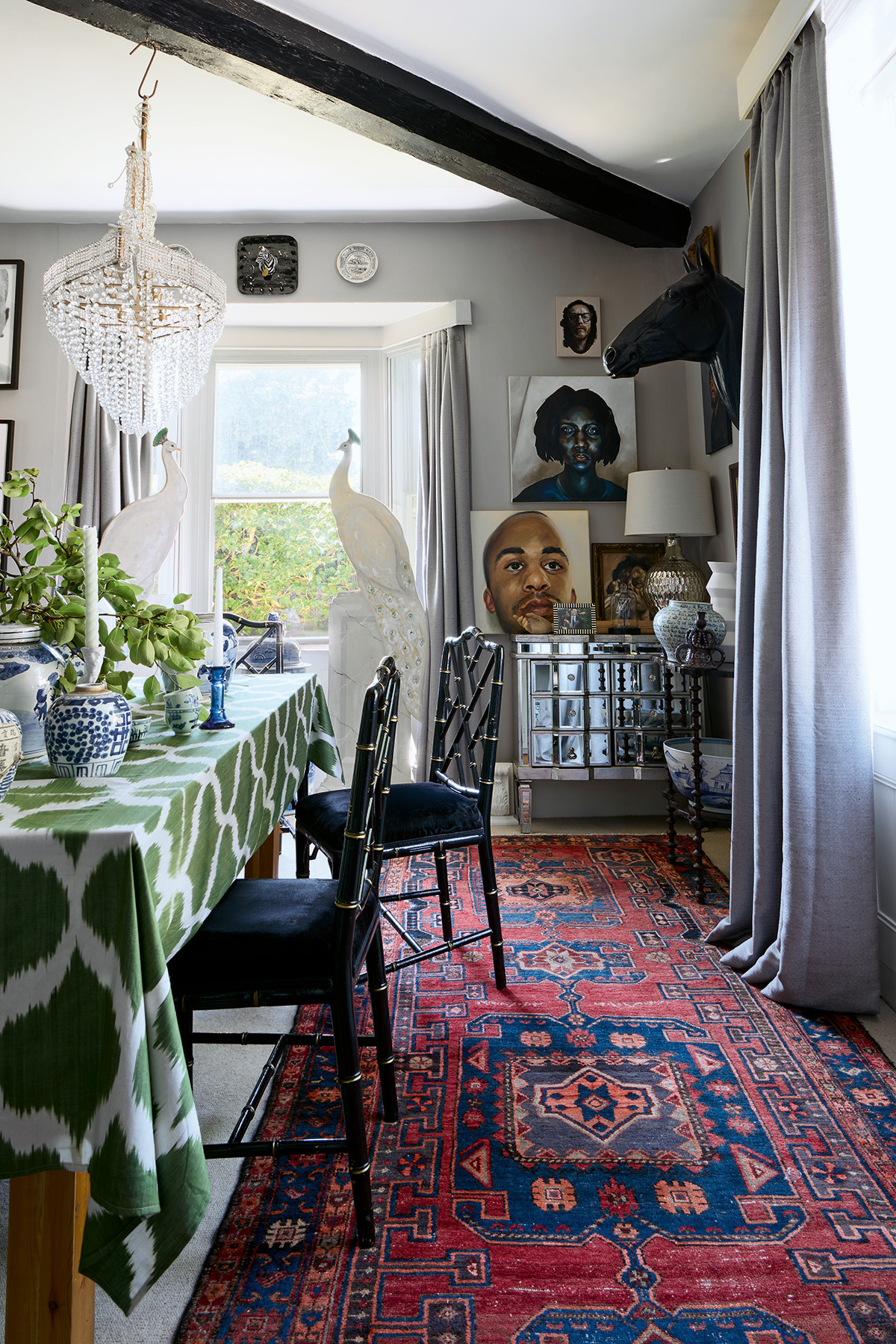 a dining room with neutral walls and curtains and a large black beam running diagonally across the ceiling. On the floor is a large persian red rug. In the centre of the room is a dining table covered with a green cloth and paired with black bamboo chairs