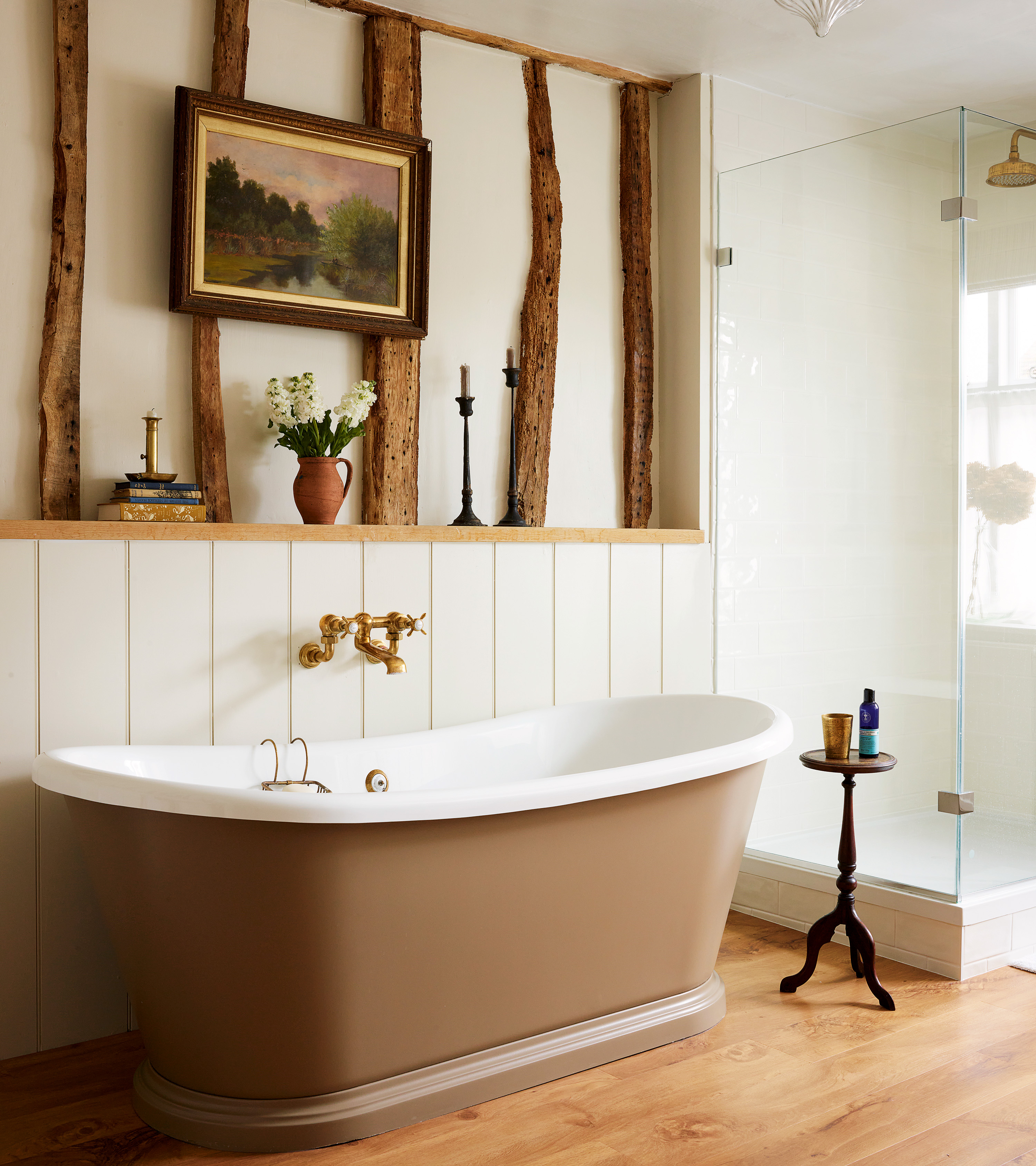 a country bathroom with a beige free standing bath tub, a glass shower enclosure and a wall behind the bath with wooden beams and a wooden ledge with toiletries on it