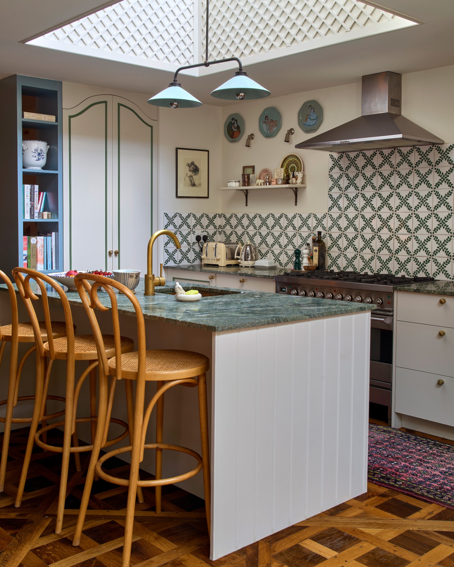 a kitchen with a versailles parquet floor, a kitchen island with a green marble top and 3 rattan bar stools tucked under it, a kitchen splashback with white and green patterned tiles and a skylight