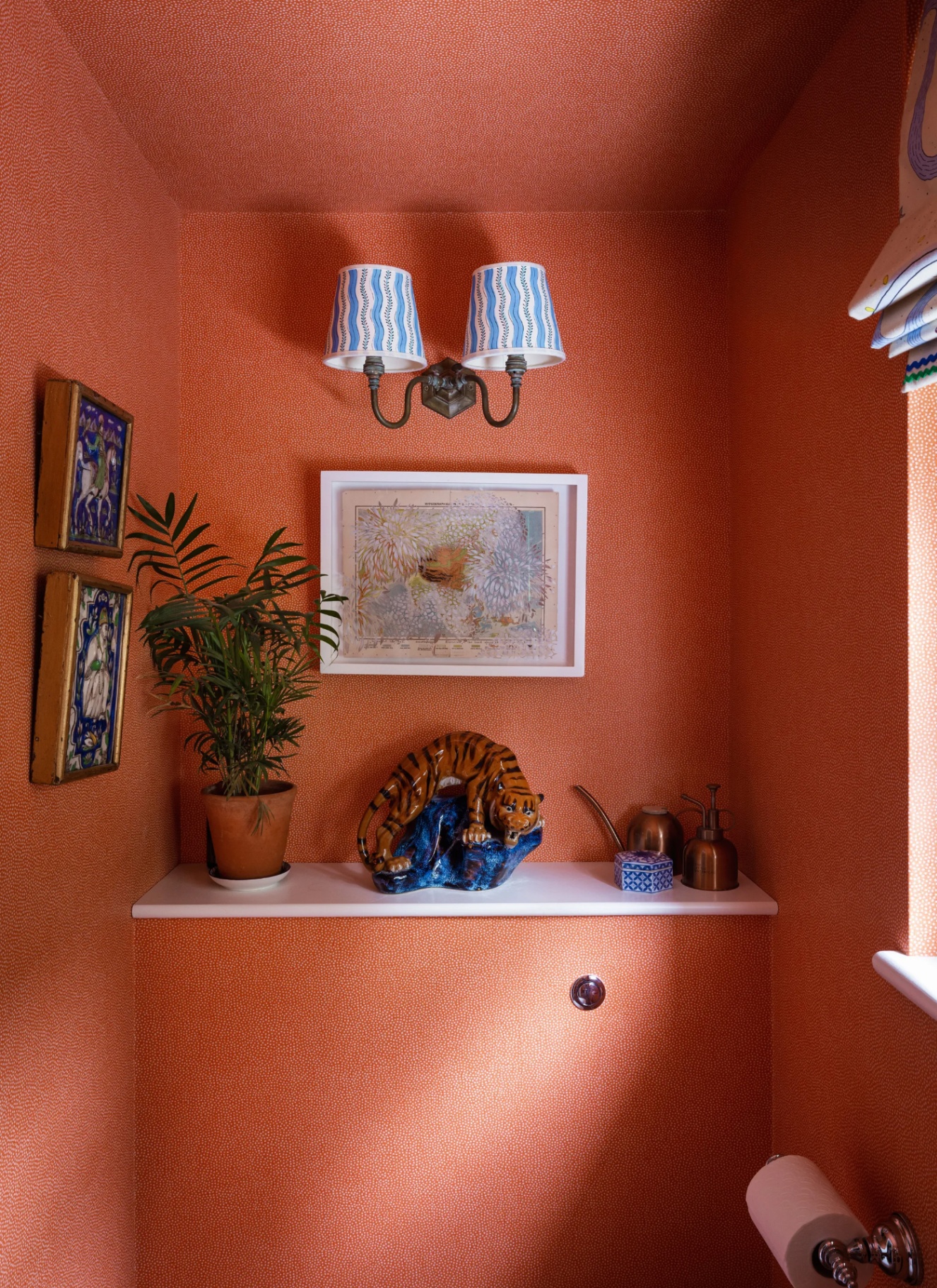 close up of a downstairs toilet wall with orange wallpaper, a ledge shelf with a plant pot, a tiger ornament and a watering can, a painting in a white frame and a double sconec with white and blue shades