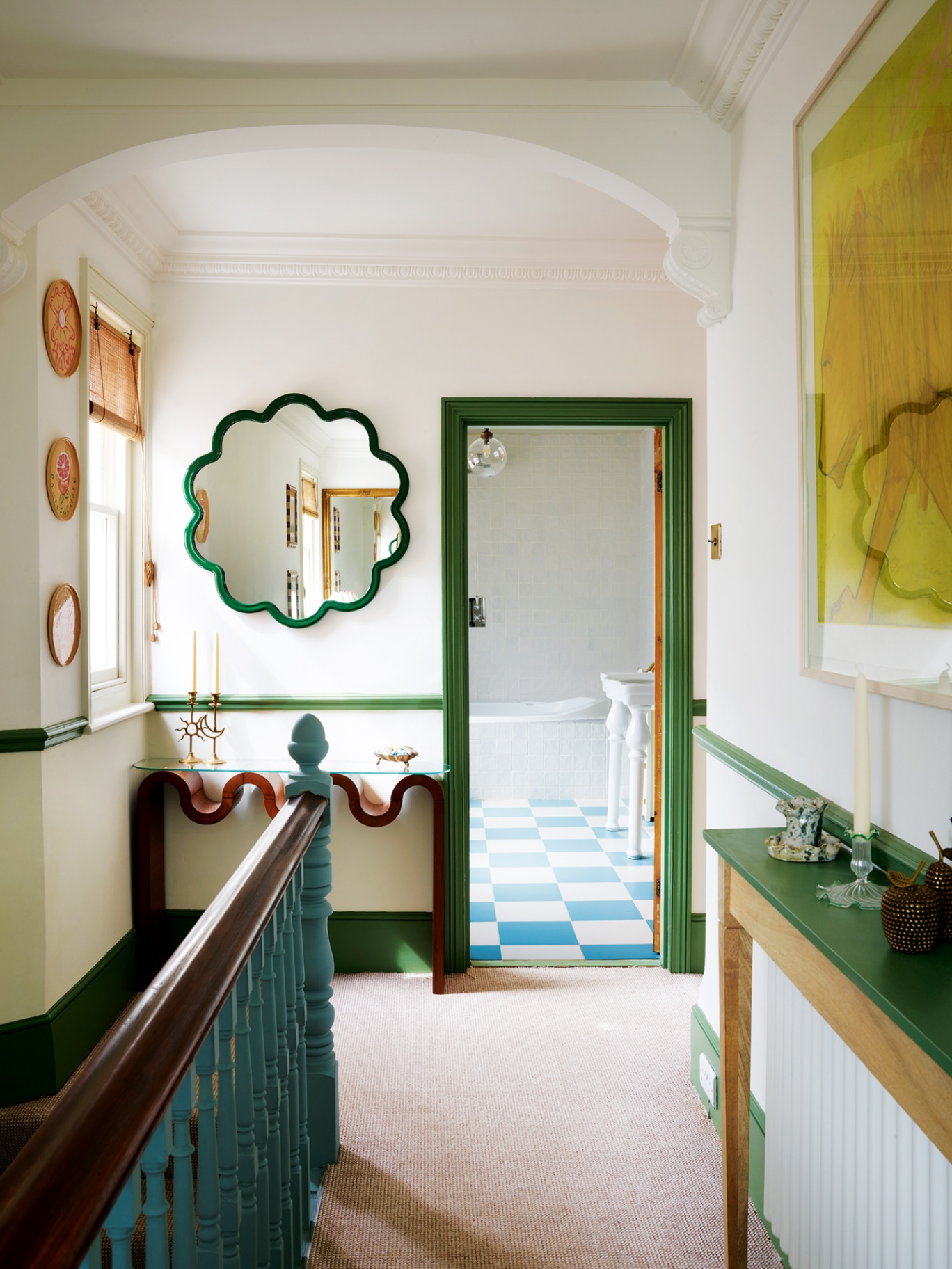 A landing of an Edwardian house leading to a bathroom seen through a door on the far wall. A wavy wooden console table with a glass top sits to the left of the door with a flower-shaped mirror above. Blue stairs appear to the left in the foreground