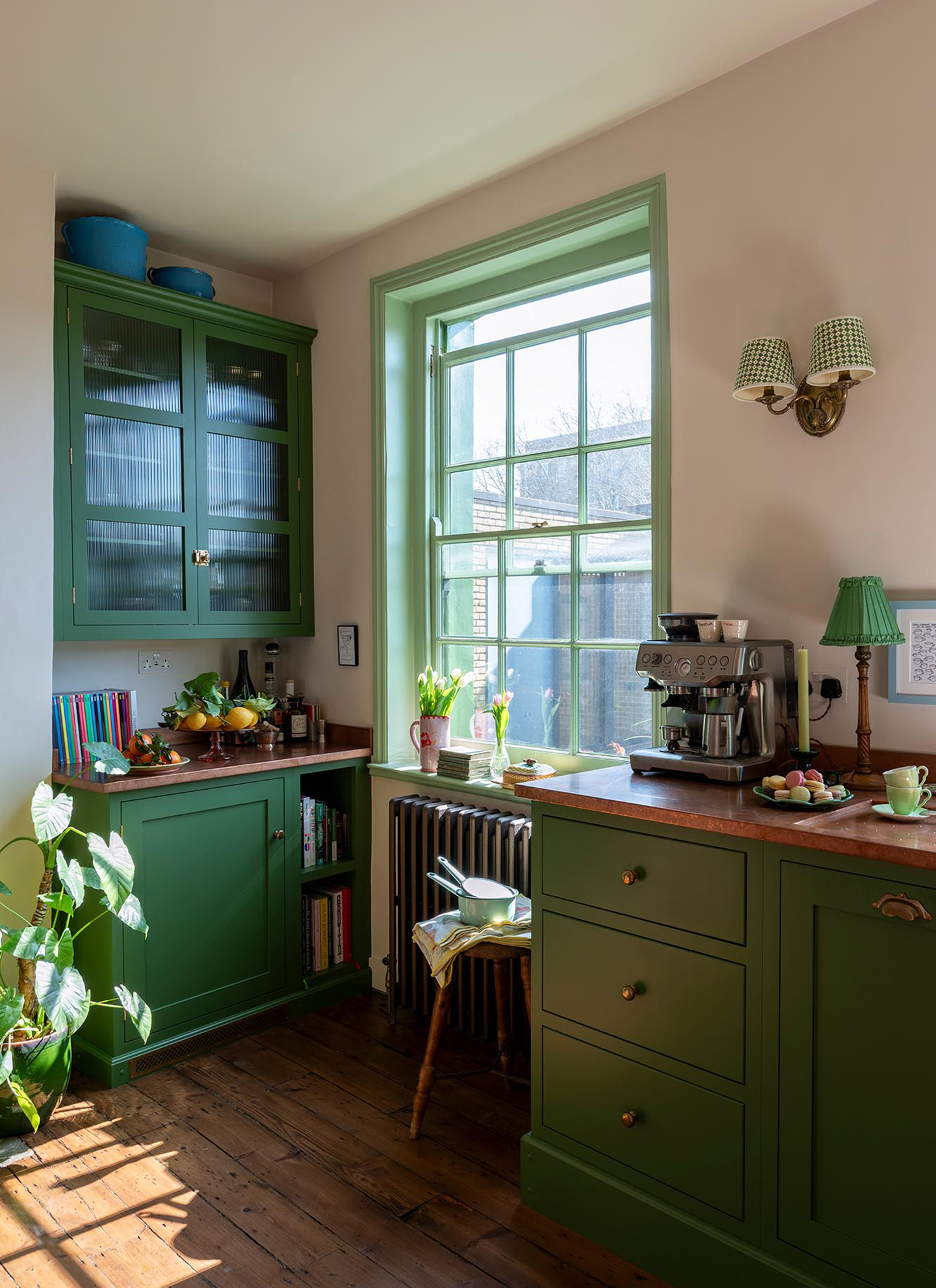 a small kitchen with bright green cabinets, pale pink walls, wooden flooring, a large sash window and a green plated wall light