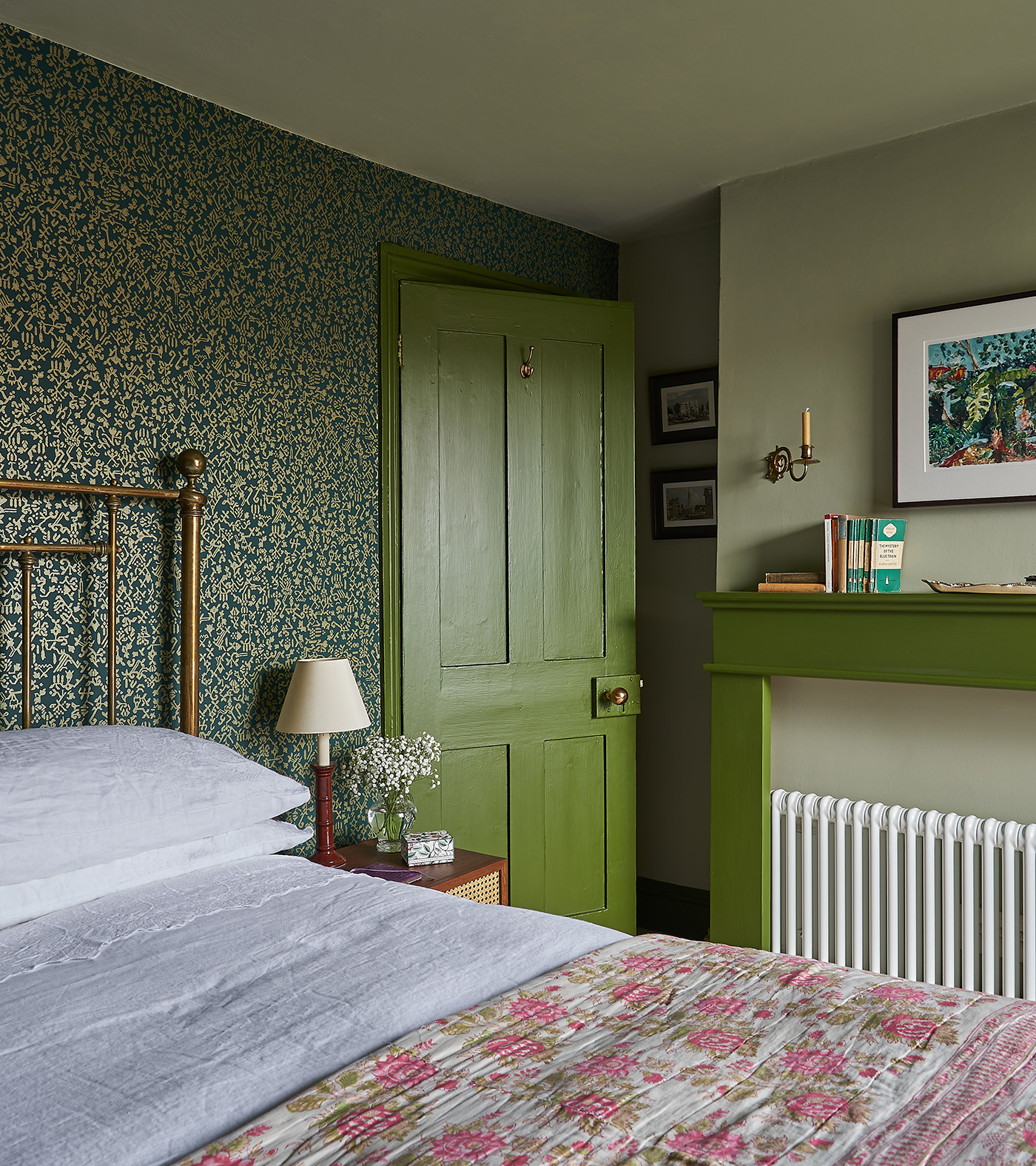 a bedroom with green botanical wallpaper, an olive green door and olive green mantel piece, with an iron bed with white bedding and a pink floral bed spread