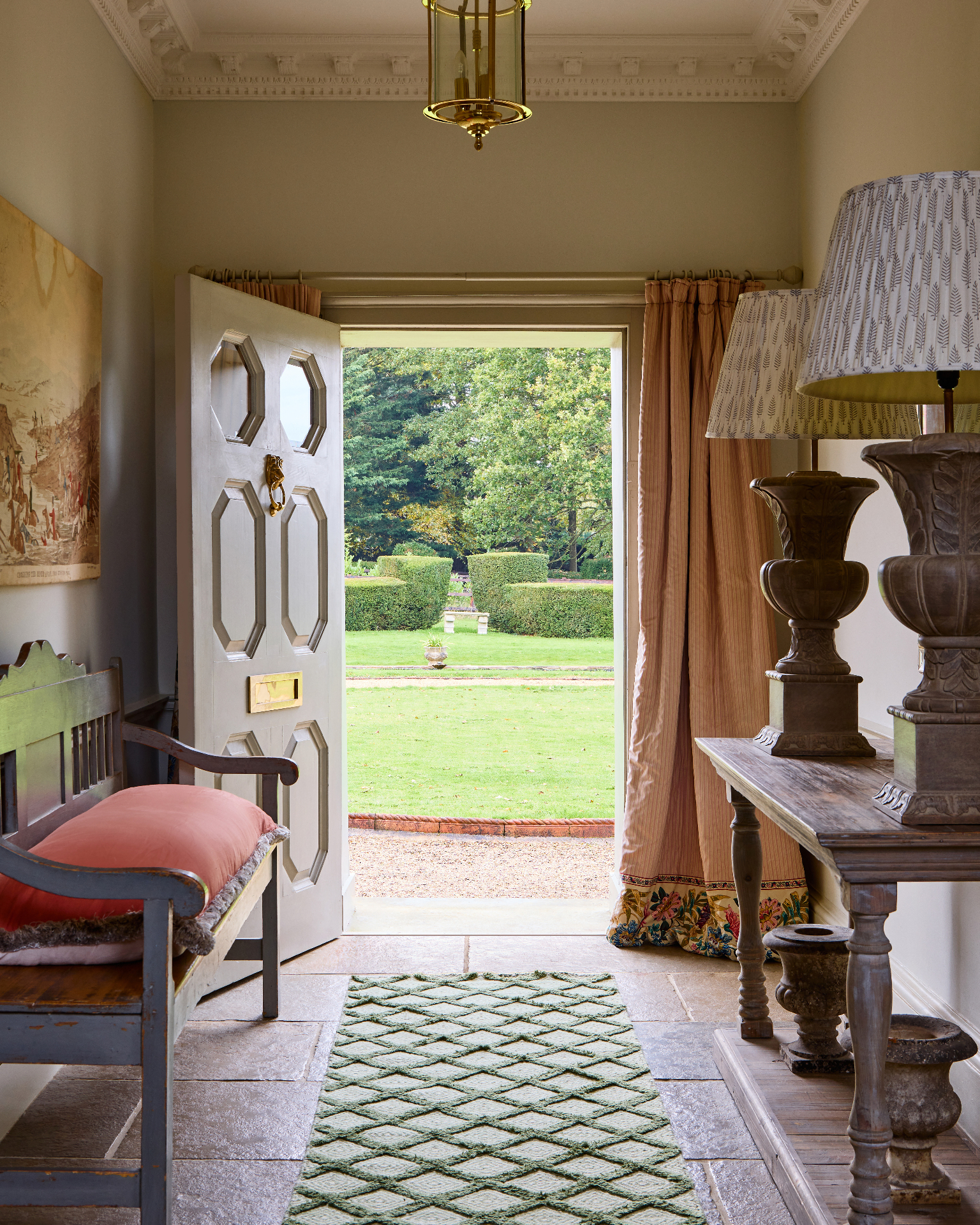A HALLWAY with an open front door looking out to a lawn, a green patterned runner, a blue painted bench and a console table with two large lamps