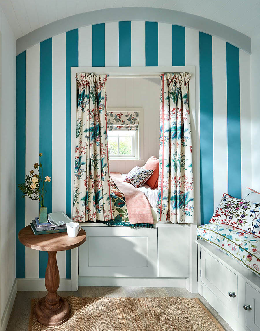 a bedroom with a bed alcove painted in a bold blue and white stripe with floral curtains, built in white drawers and a wooden side table