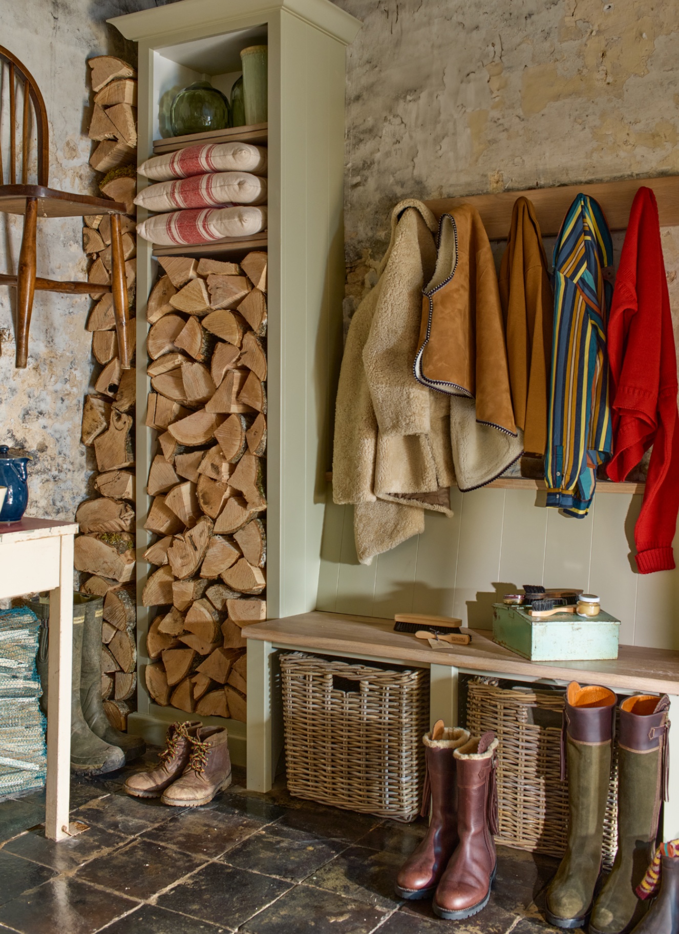 a boot room corner with black flagstone flooring, a tall open cupboard filled with logs, a bench with wicker backets under it and pegs with coats