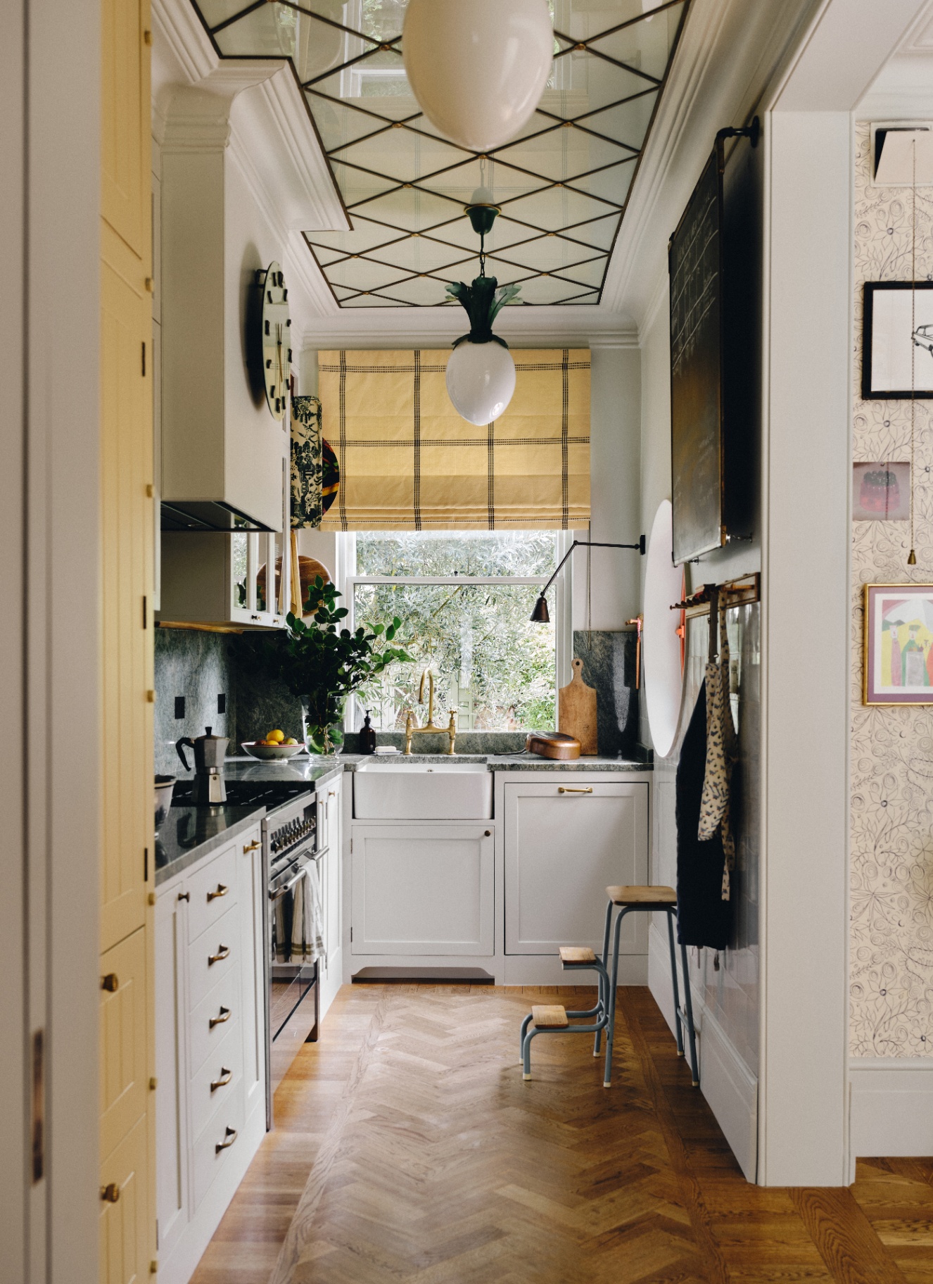 a narrow kitchen with white cabinets, wooden flooring, a window with a pale yellow blind and two large egg shaped pendant lights