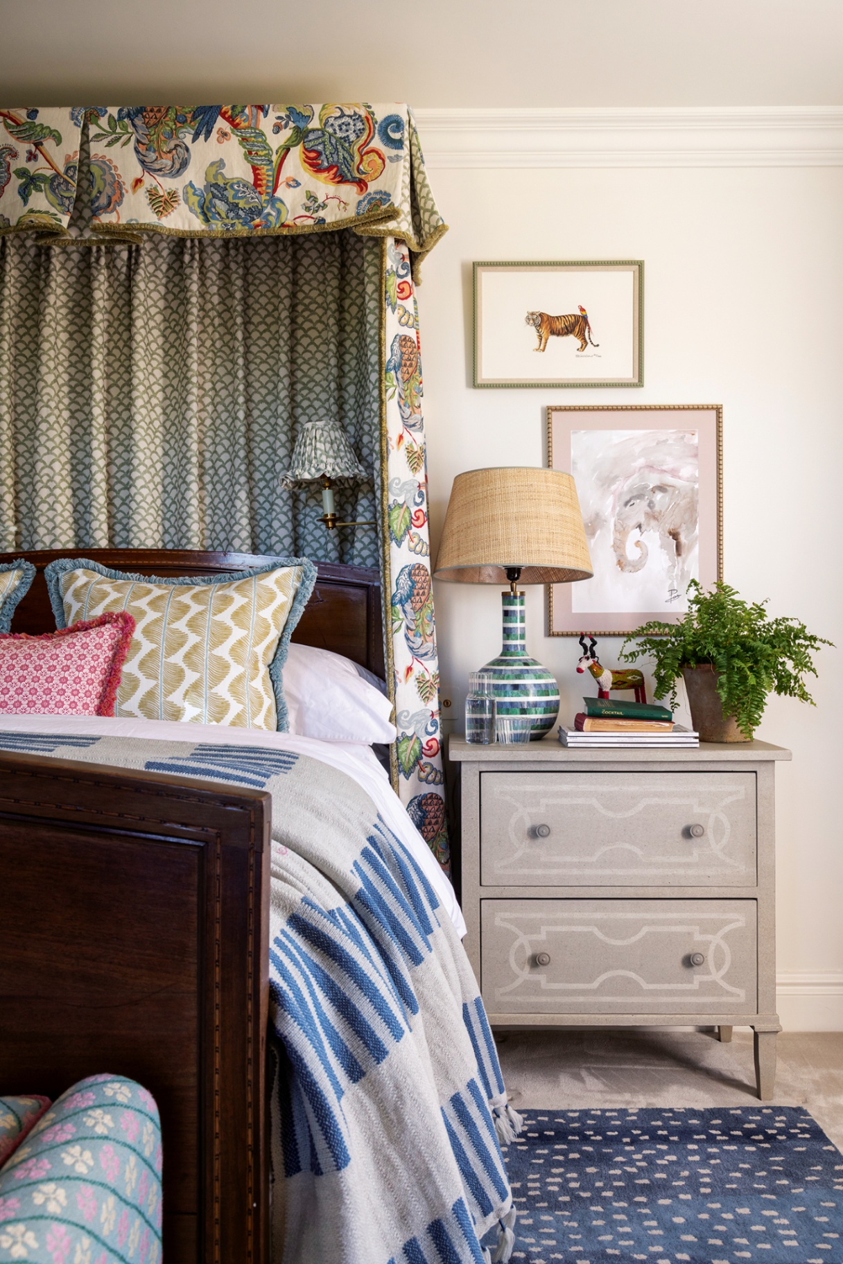 A bedroom with a large wooden bed in a blue and white spotty rug with a grey bedside table next to it, topped with a table lamp, plant and books. A floral half-tester hangs above, with a small wall light positioned above the headboard