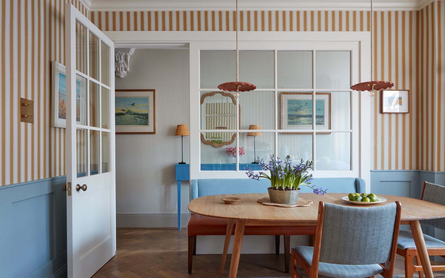 a dining room with yellow and white striped wallpaper on half of the wall and blue panelling on the bottom half, a large wooden oval dining table with blue upholstered chairs and 2 pendant lights above it
