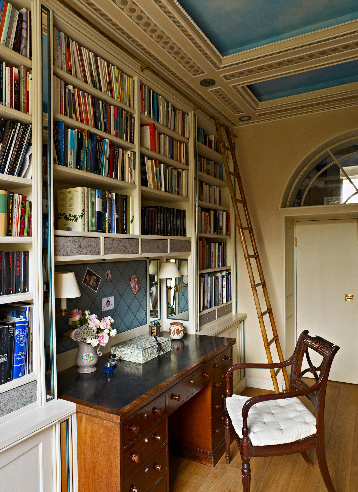 a study with a dark wood desk and a matching chair, a library wall full of books with a ladder and a ceiling painted in a blue sky effect