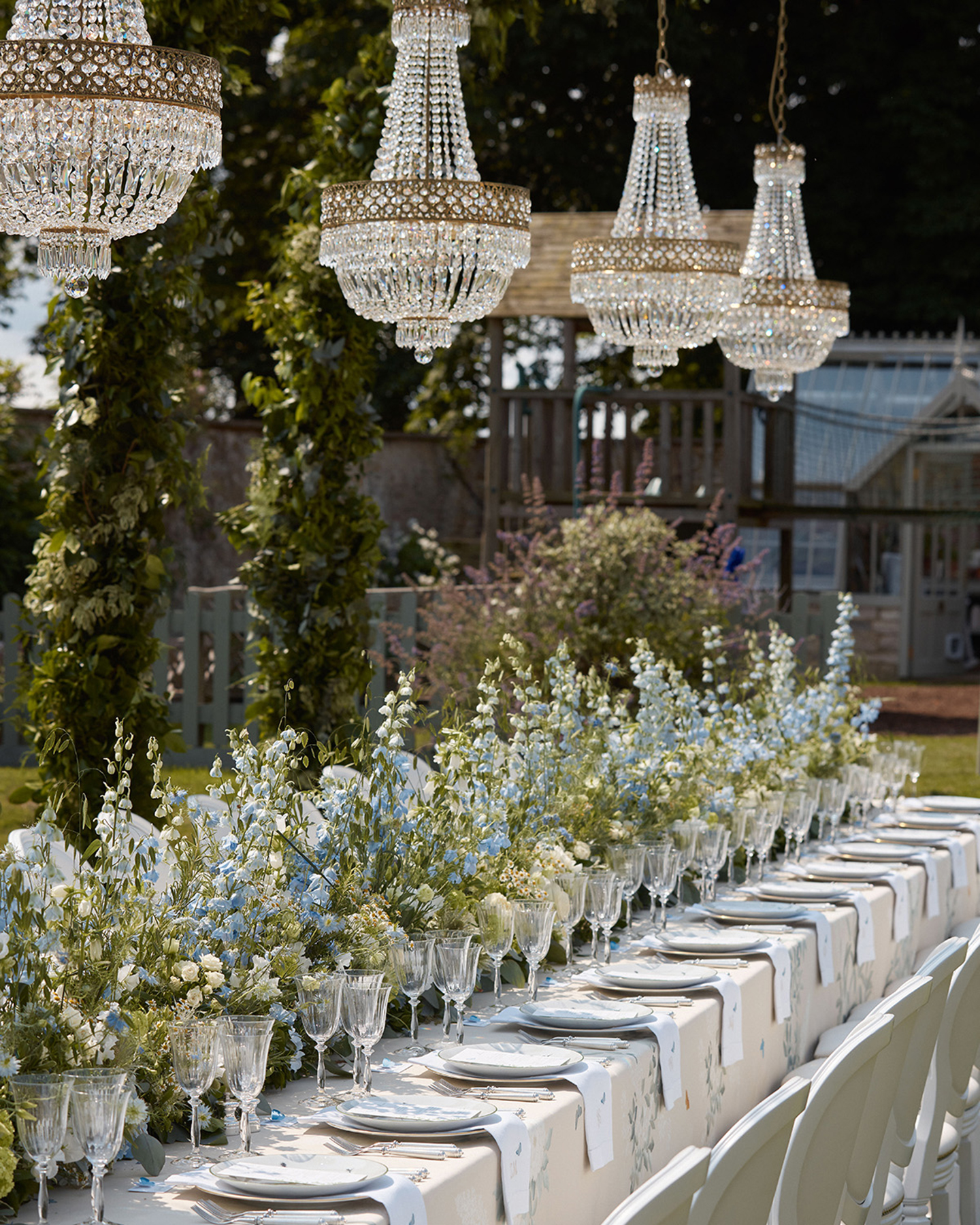 an outdoor garden party table with flowers down the centre of the table, a row of plates, wine glasses and napkins with hanging crystal chandeliers above