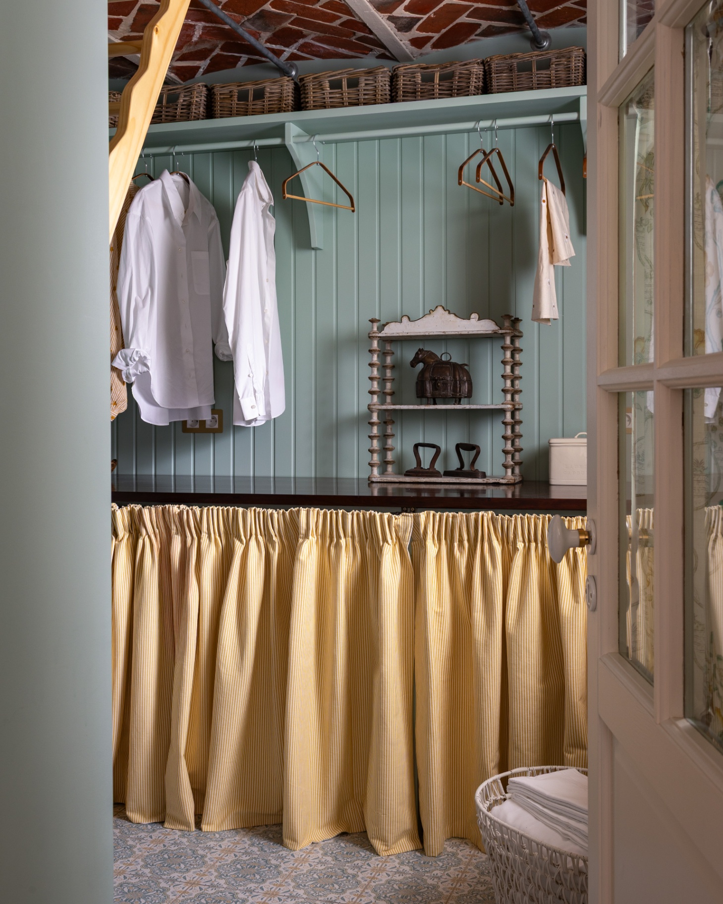 a utility room with sage green panelled walls, a long fabric skirt covering a cupboard and a hanging rail with clothes and coat hangers