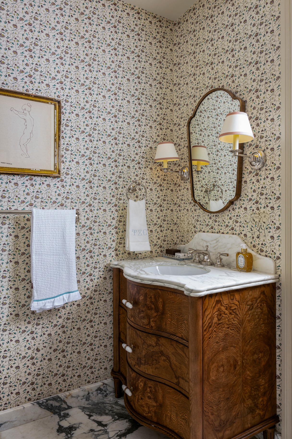 A small bathroom with a wooden vanity with a marble top, dainty floral wallpaper and marble flooring. A mirror hangs above the basin, flanked by two small wall lights, with a hand towel, larger towel rail and artwork hung beside it 