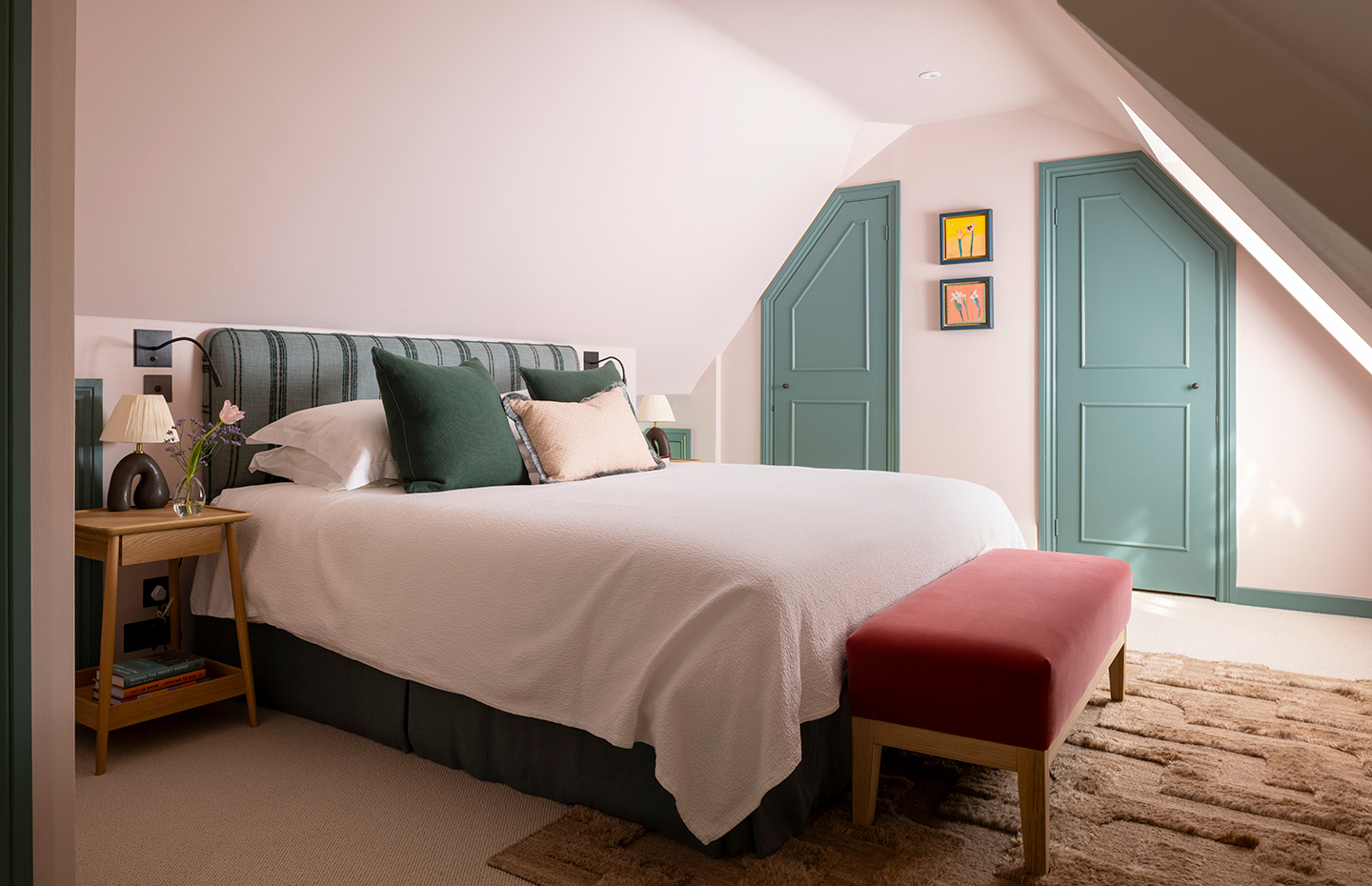 a bedroom with a sloped ceiling, with walls painted in a light pink and skirting boards and two doors painted in green. A double bed is dressed in white linen and green cushions with a pink bench at the end and paired with a wooden bedside table