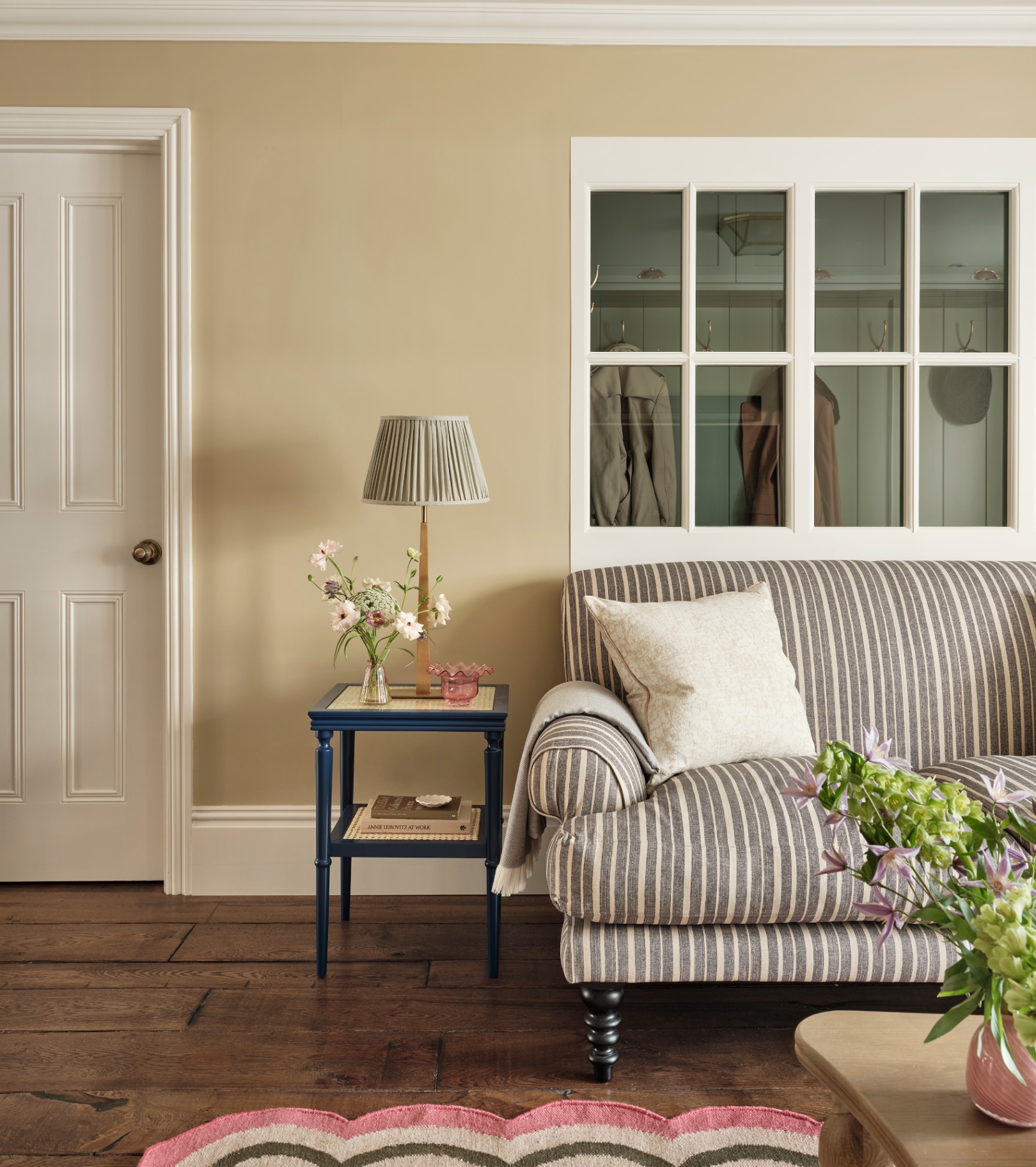 a living room corner with dark cream beige painted walls, a striped sofa in front of an internal window, and a navy and rattan side table with a lamp on next to the sofa