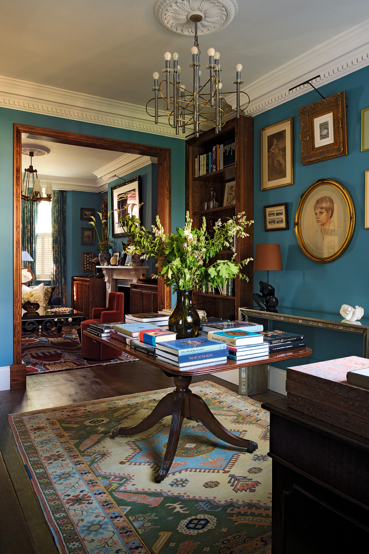 a reception room with a rectangular wooden table in the middle, on a patterned vintage rug, topped with flowers in a vase and stacks of books. The walls are painted deep blue and hung with various artworks. A doorway looks through to the living room behind