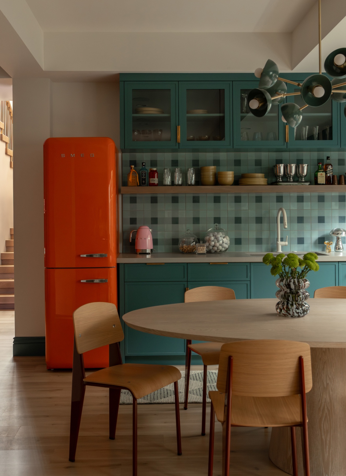 Open plan kitchen dining area with a oval wooden table, Scandinavian wooden chairs, teal cabinets, and a bright orange retro fridge
