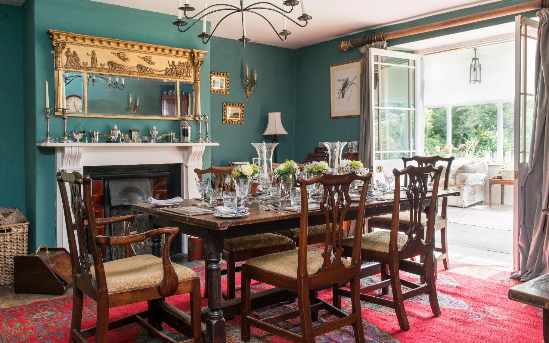 Teal green dining room with a long wooden table, traditional carved chairs, a red patterned rug, white fireplace with an ornate gold mirror, metal chandelier, and open French doors leading to a garden room