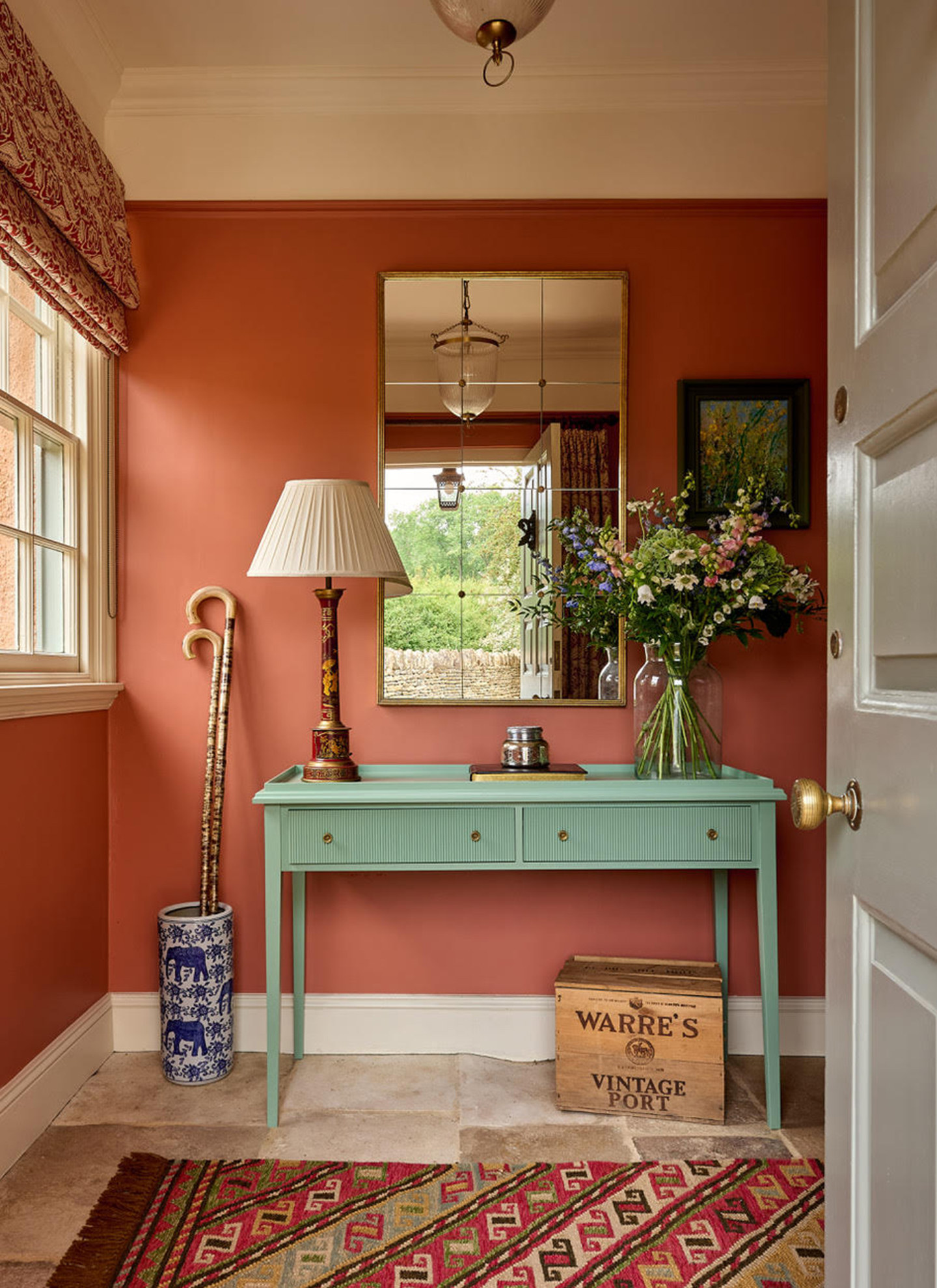 a hallway with terracotta walls, a mint green console table with a vase of fresh flowers and a panelled mirror above it