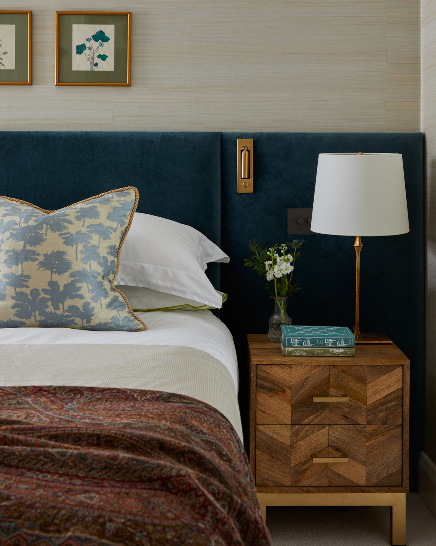 close up of a bedroom corner with neutral textured wallpaper, a wooden side table with a white lamp, a gold reading light attached to a navy velvet headboard and a bed with white bedding