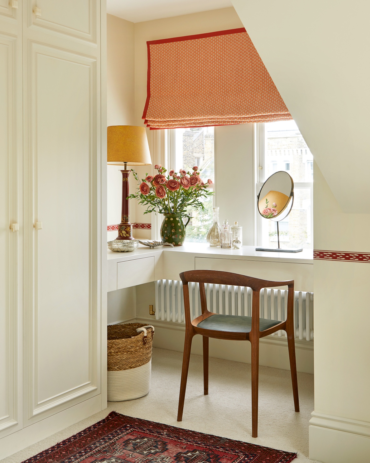 a corner of a room with a dressing table and chair facing the window, an orange blind and a red persian style runner on the floor