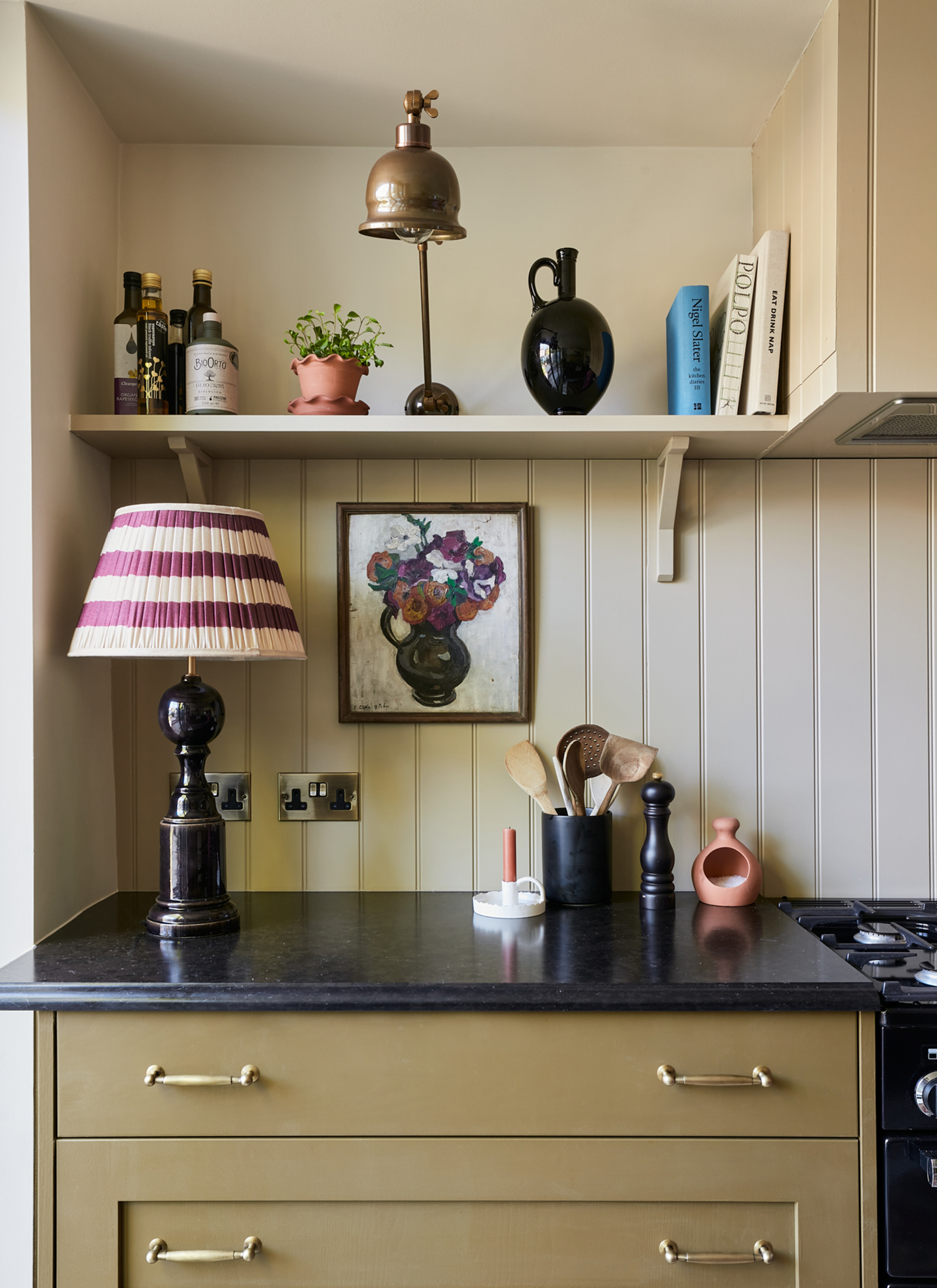 a kitchen corner with olive cabinetry, black countertops, cream tongue and groove panelling and an open shelf with bottles and books