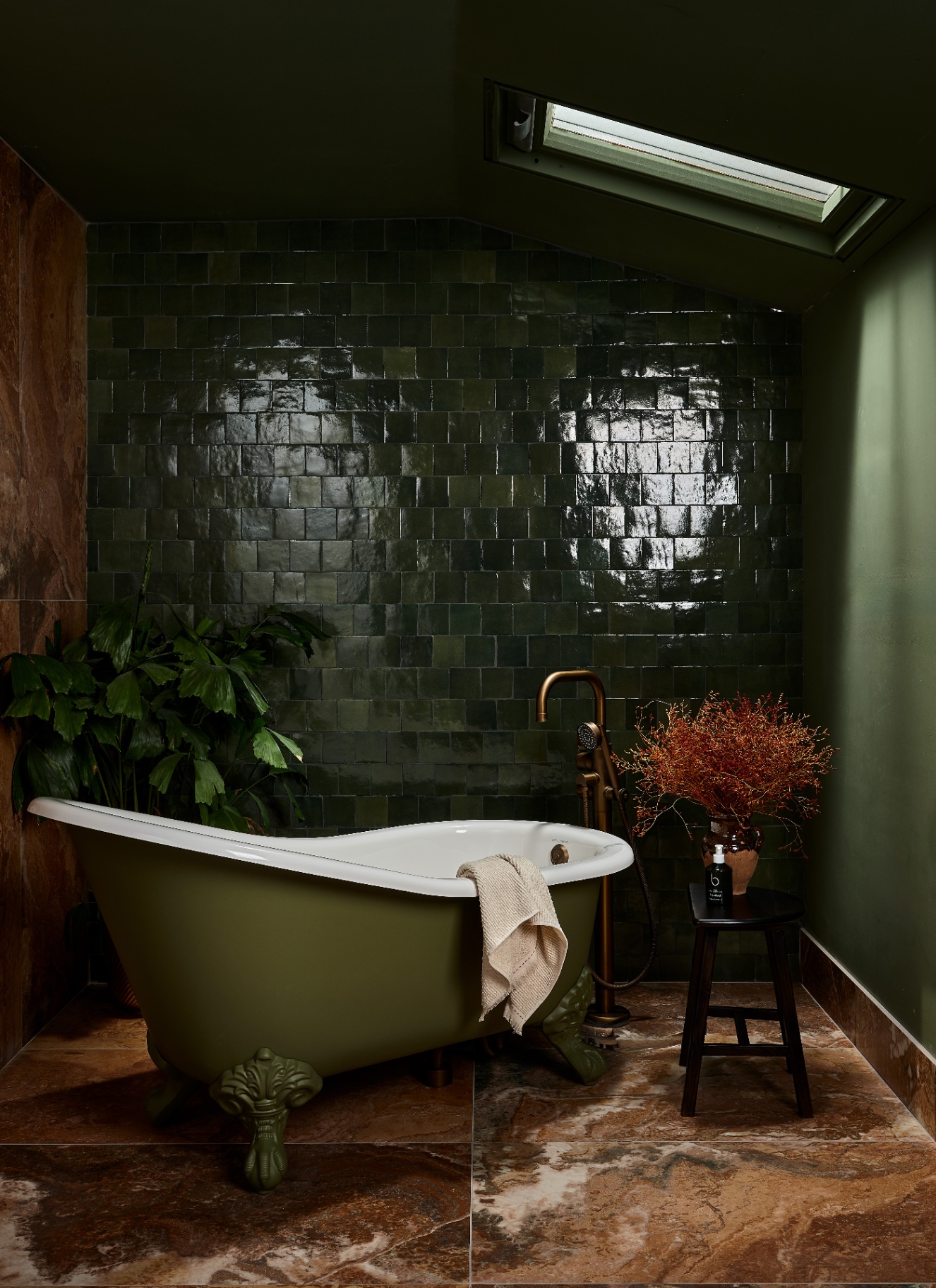 Forest green bathroom with an olive green clawfoot tub set against glossy dark green square wall tiles and reddish-brown marble floor tiles. Natural light streams from a ceiling skylight onto a small wooden stool with dried foliage and a large leafy plant