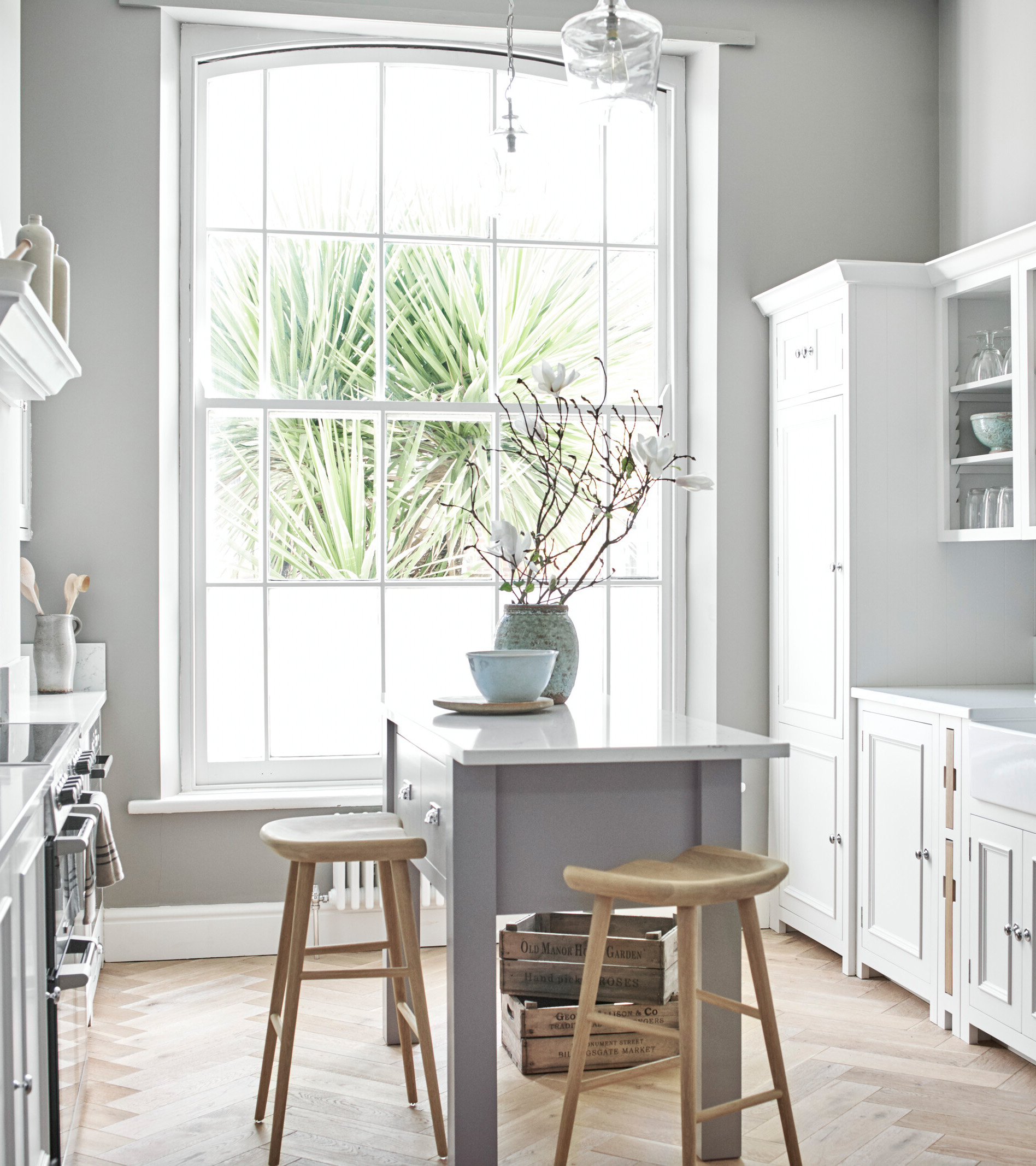 a kitchen with a central island painted grey with white marble top, two wooden stools next to it and a vase on top. White cabinetry and brown wooden parquet floor