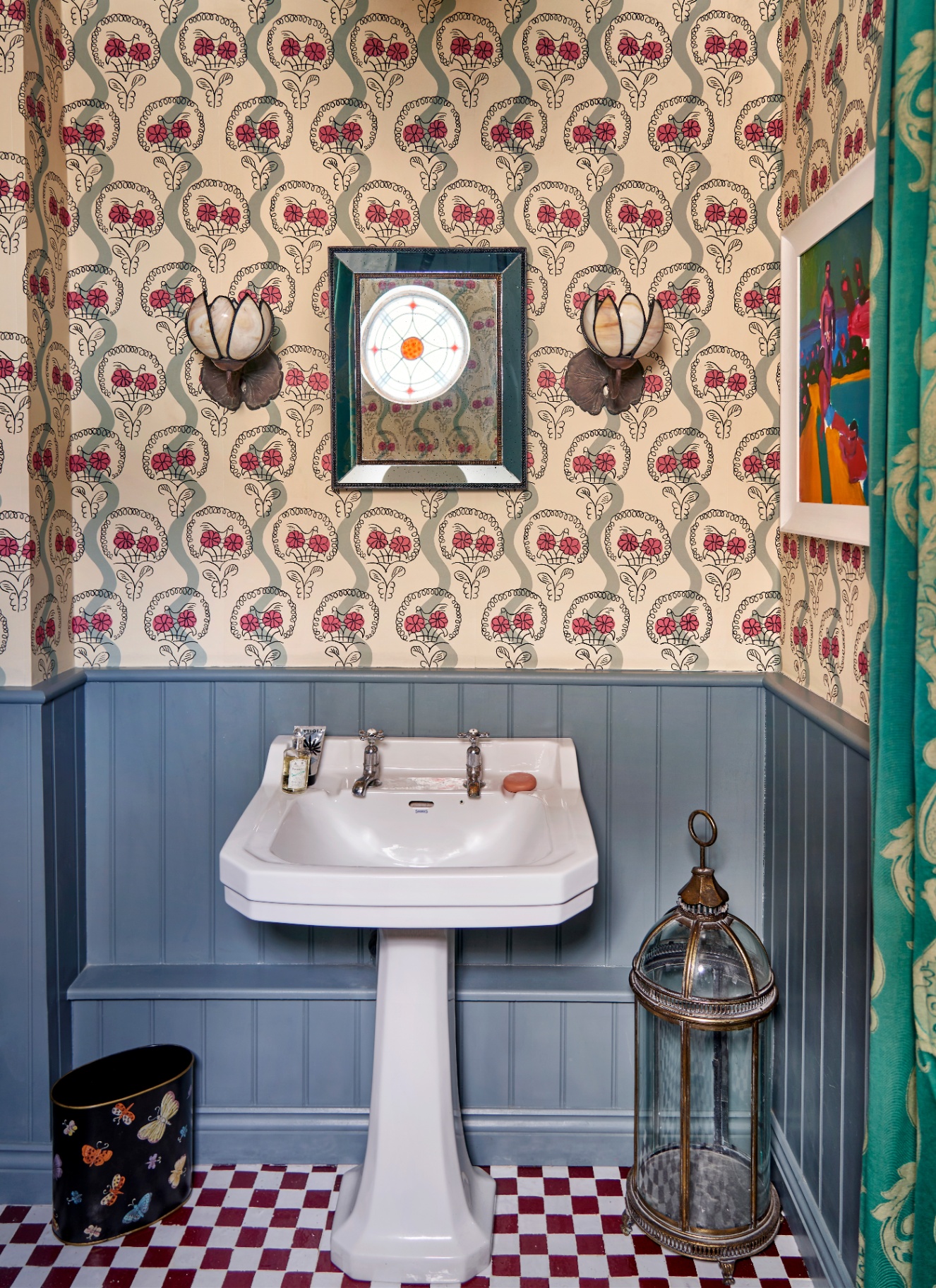 a downstairs toilet with half pale blue panelled walls and half cream and pink floral wallpaper, a white sink, red and white checkerboard flooring, a large lantern on the floor, a small mirror and two flower shaped sconces