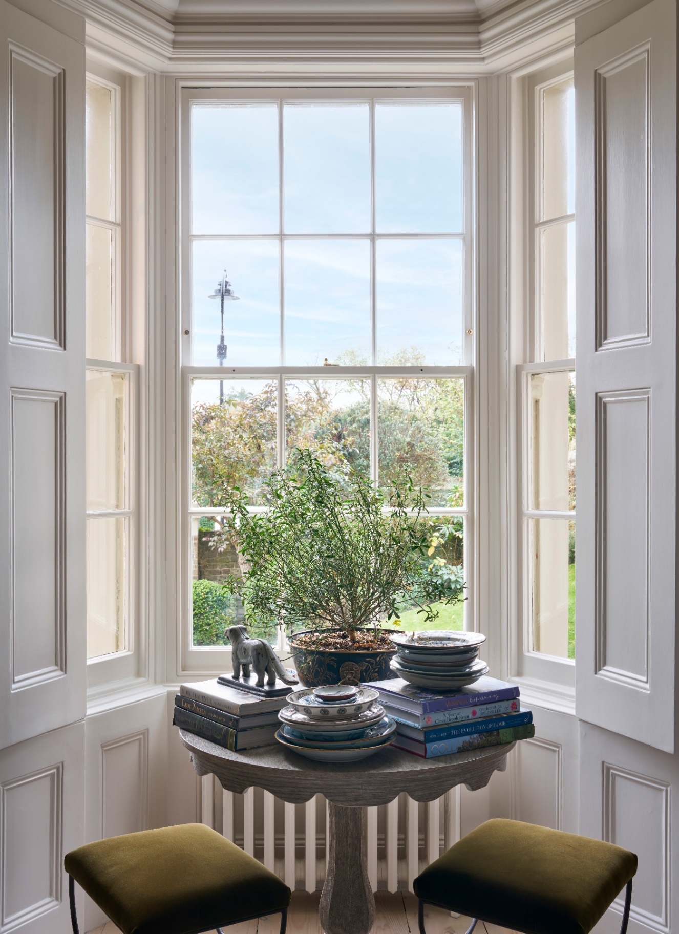a large sash window with a round table in front of it with piles of books on top and two olive green velvet stools