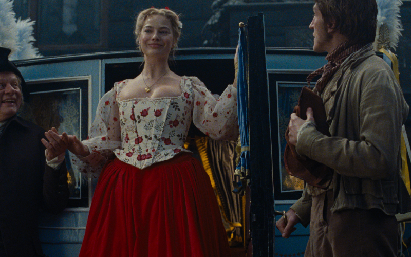 Margot Robbie as Cathy in Wuthering Heights - a woman with blonde hair steps out of a blue carriage wearing a red and white victorian dress