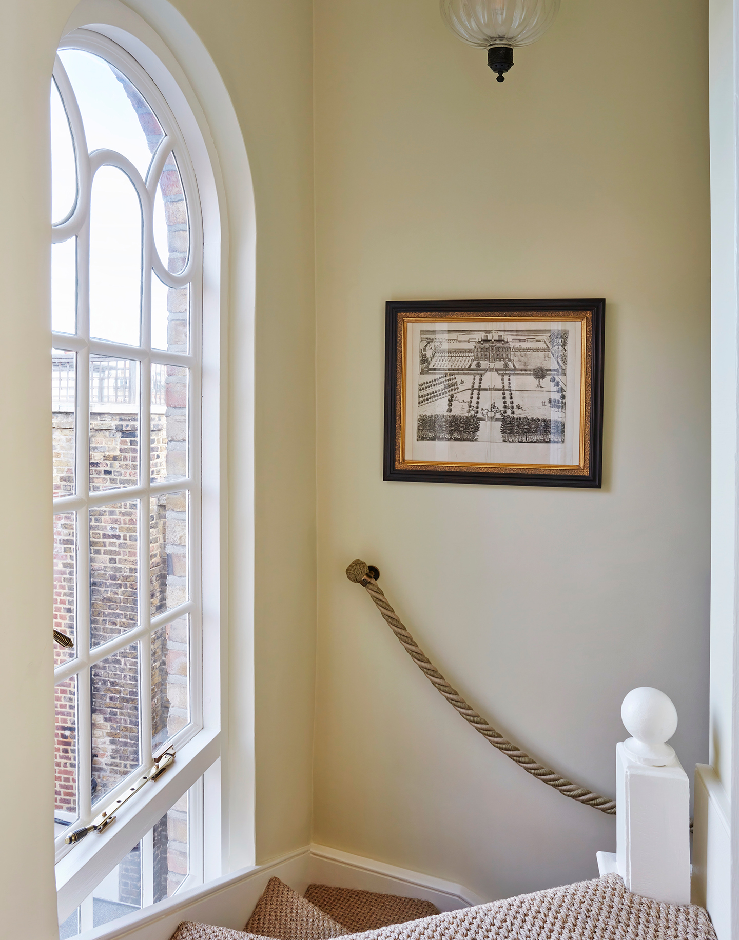 A curved stairway with a rope handrail, an antique artwork hanging on the wall and, to the left, a large arched period window