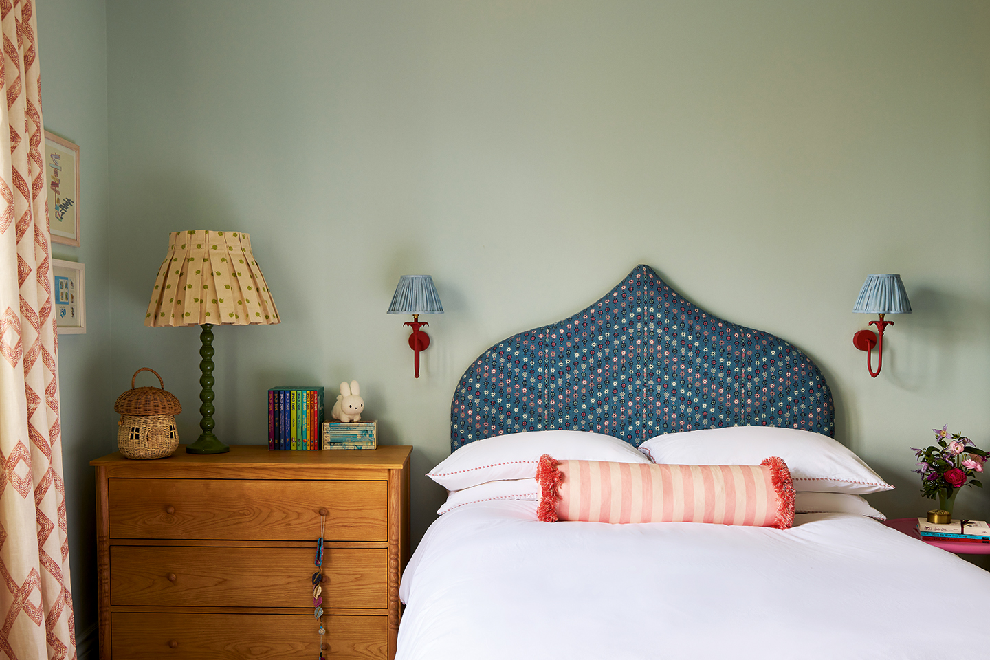 A close up of a childrens room with blue walls, an ogee shaped patterned blue headboard, wooden chest of drawers topped with a bobbin lamp and pleated shade, and red wall lights topped with blue shades. To the left is the edge of red and white curtains