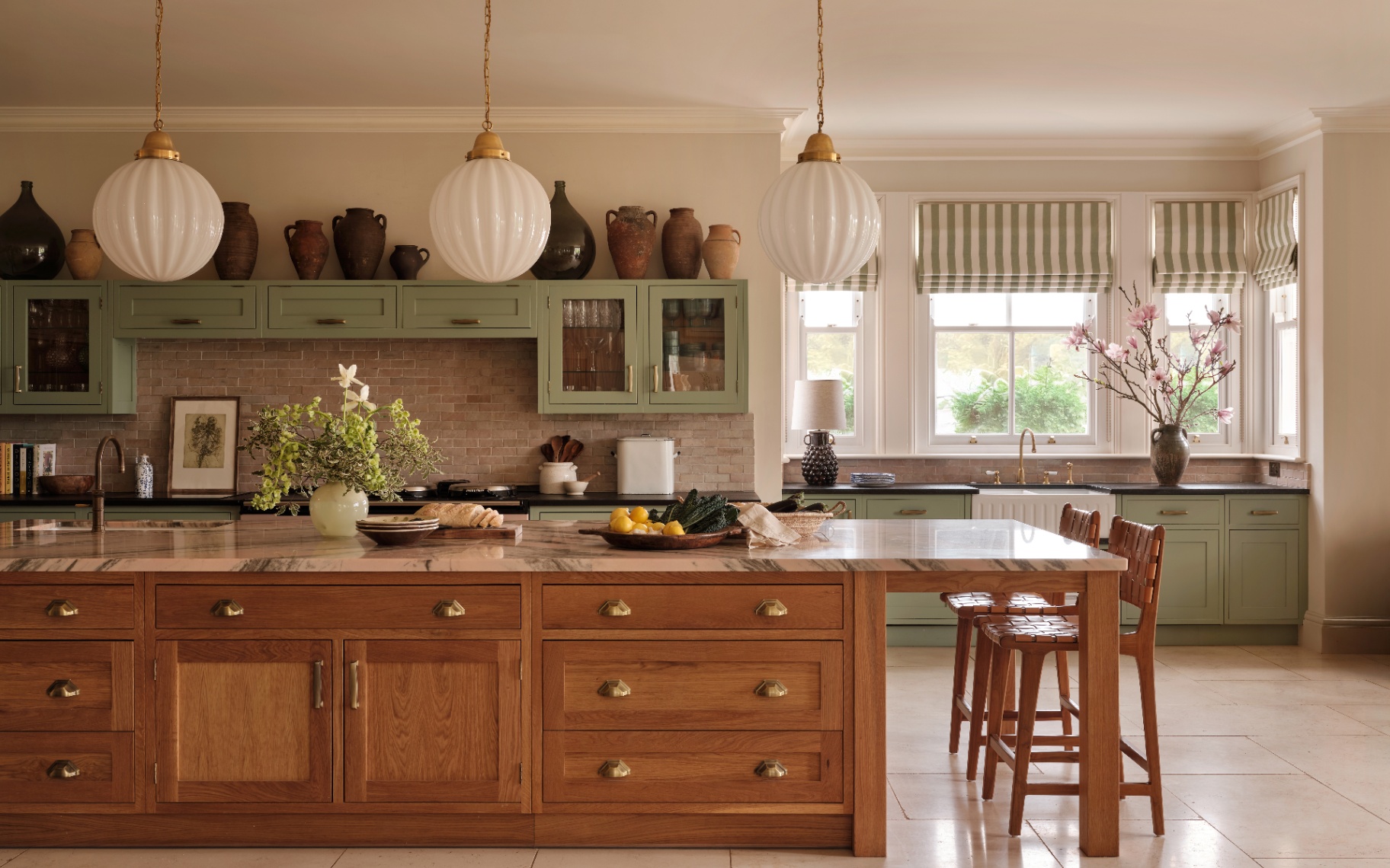 a kitchen with 3 large statement sphere pendant lights hanging above a long wooden kitchen island with a white and grey marble top and two woven leather dining chairs tucked underneath
