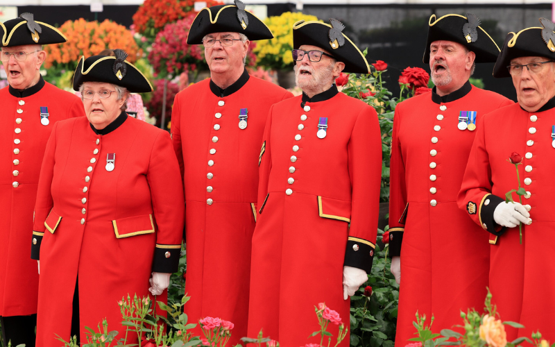 The Chelsea Pensioners singing at the Chelsea Flower Show in 2024, they are white men with grey hair wearing their red uniforms and black tricorne hats