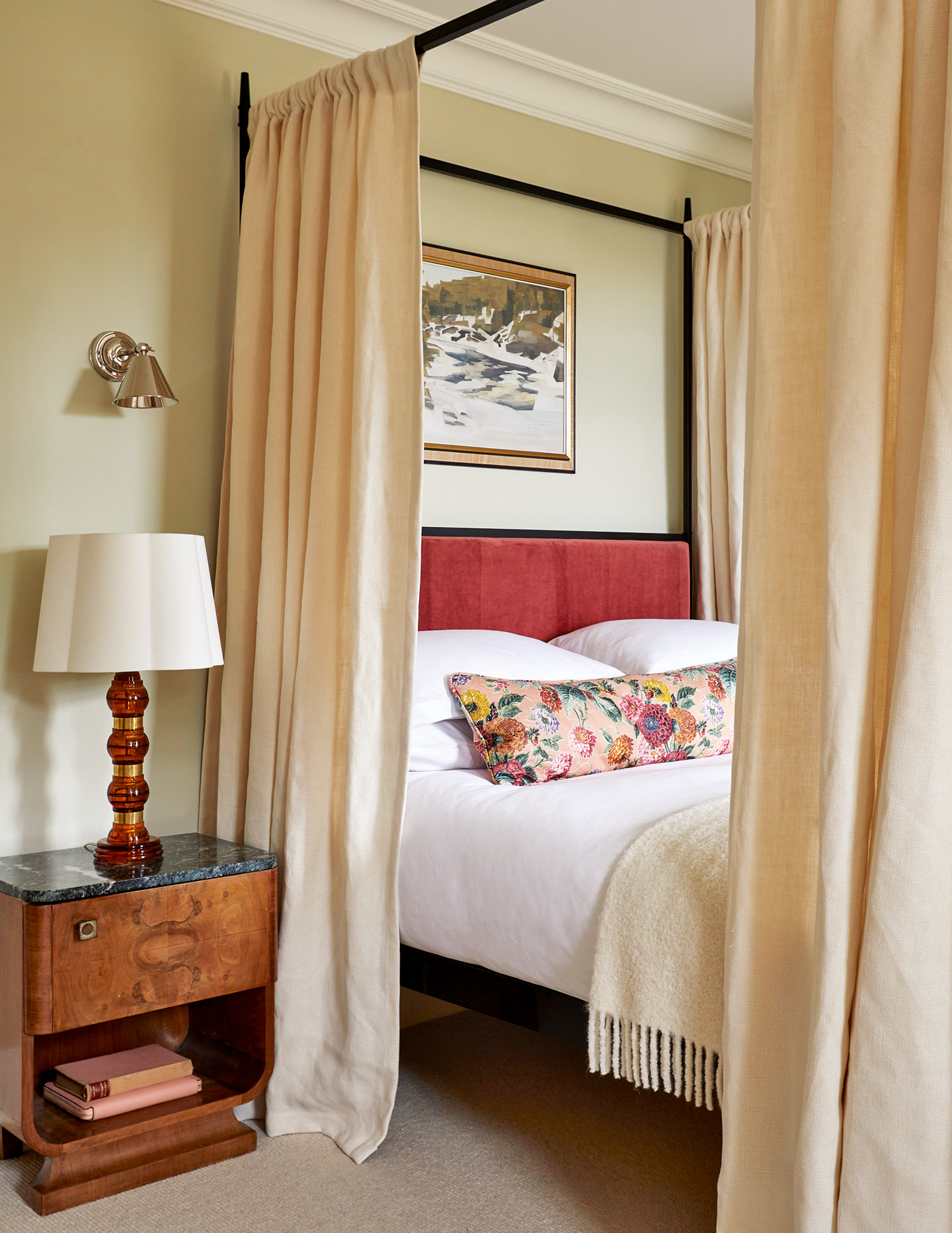 A light green bedroom with a metal four-poster bed with neutral linen curtains. The bed has a red headboard and is topped with a floral cushion and wool blanket. A burl wood bedside table with marble top holds a glass lampshade and some books