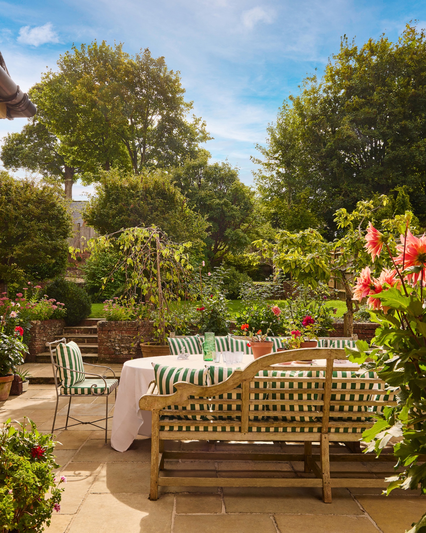 a garden patio with a large outdoor dining set with two long benches and a chair with white and green striped cushions surrounded by trees and flowers