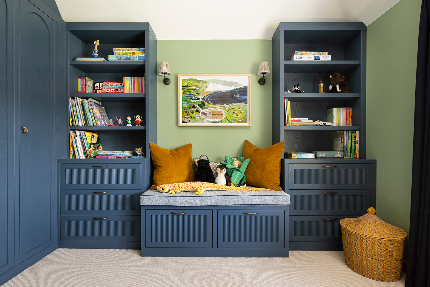 a childrens playroom with dark blue built in bookcases, wardrobes and bench seating, topped with stuffed toys and cushions. Above is a colourful countryside-themed artwork with wall lights either side and on the floor is a striped wicker storage basket