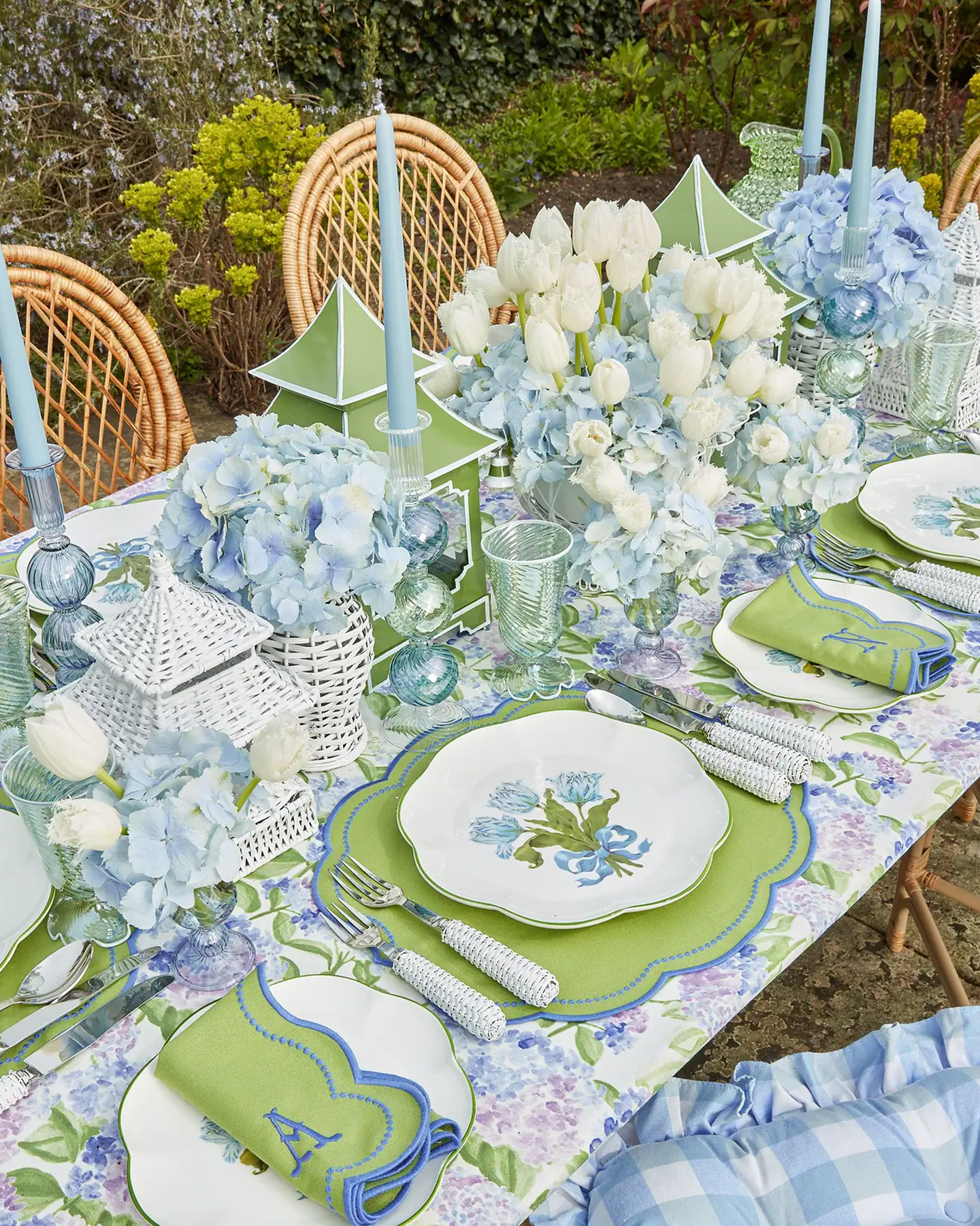an outdoor tablescape with white and blue flowers down the centre, green and blue placemats, white plates with blue floral motifs and personalised initial green and blue napkins